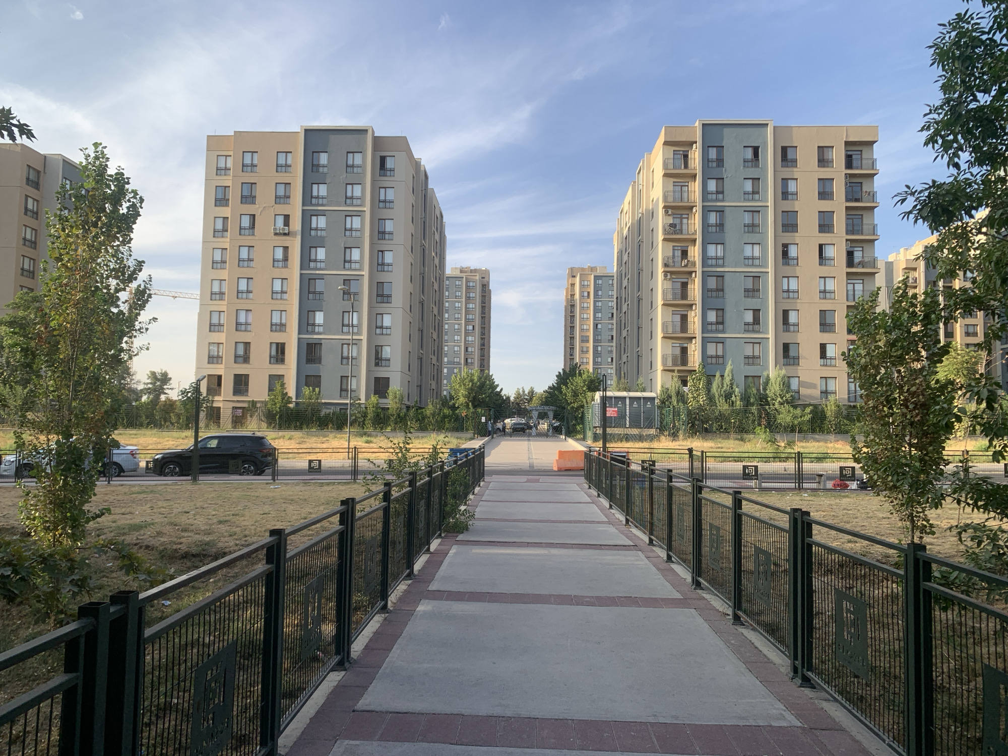 An outdoor, eye-level, wide shot captures a pedestrian walkway leading towards apartment buildings under a partly cloudy sky. The walkway is paved with concrete slabs bordered by red bricks, with a black metal fence lining both sides. Beyond the fence, dry grass and small trees are visible.
On the left side of the image, a tall, multi-story apartment building with beige and light blue facades stands partially obscured by a leafy tree. A construction crane can be seen in the distance behind the building. Further to the left, another apartment building with similar color schemes is visible. A black SUV is parked in the background on the left, with a white car parked in front of it.
On the right side of the image, a similar tall apartment building with beige and light blue facades is featured, also with balconies. More trees and vegetation are present in the foreground and midground.
In the distance, several more apartment buildings of varying heights and colors are visible, with a few trees interspersed between them. The walkway appears to lead into a complex or neighborhood with more buildings and a gateway structure in the far distance. The overall scene suggests a residential area with modern architecture.