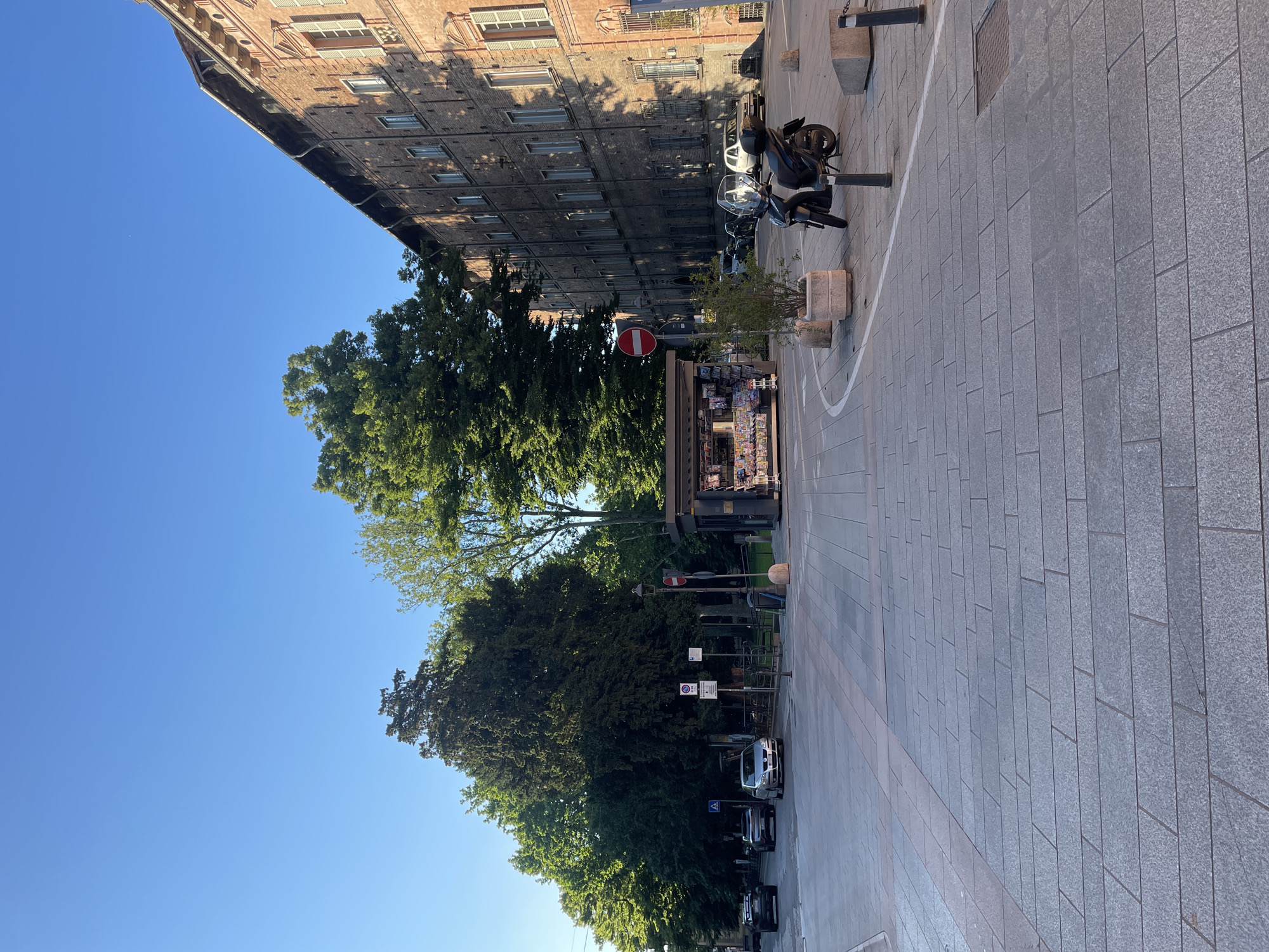 The image shows a street scene in Piacenza, Italy. A multi-story brick building with closed shutters lines the left side of the street. In front of the building is a paved pedestrian zone with trees, street signs, and parked cars. A newsstand stands along the pedestrian area. Parked motorbikes are also visible. The sky is clear and blue.
