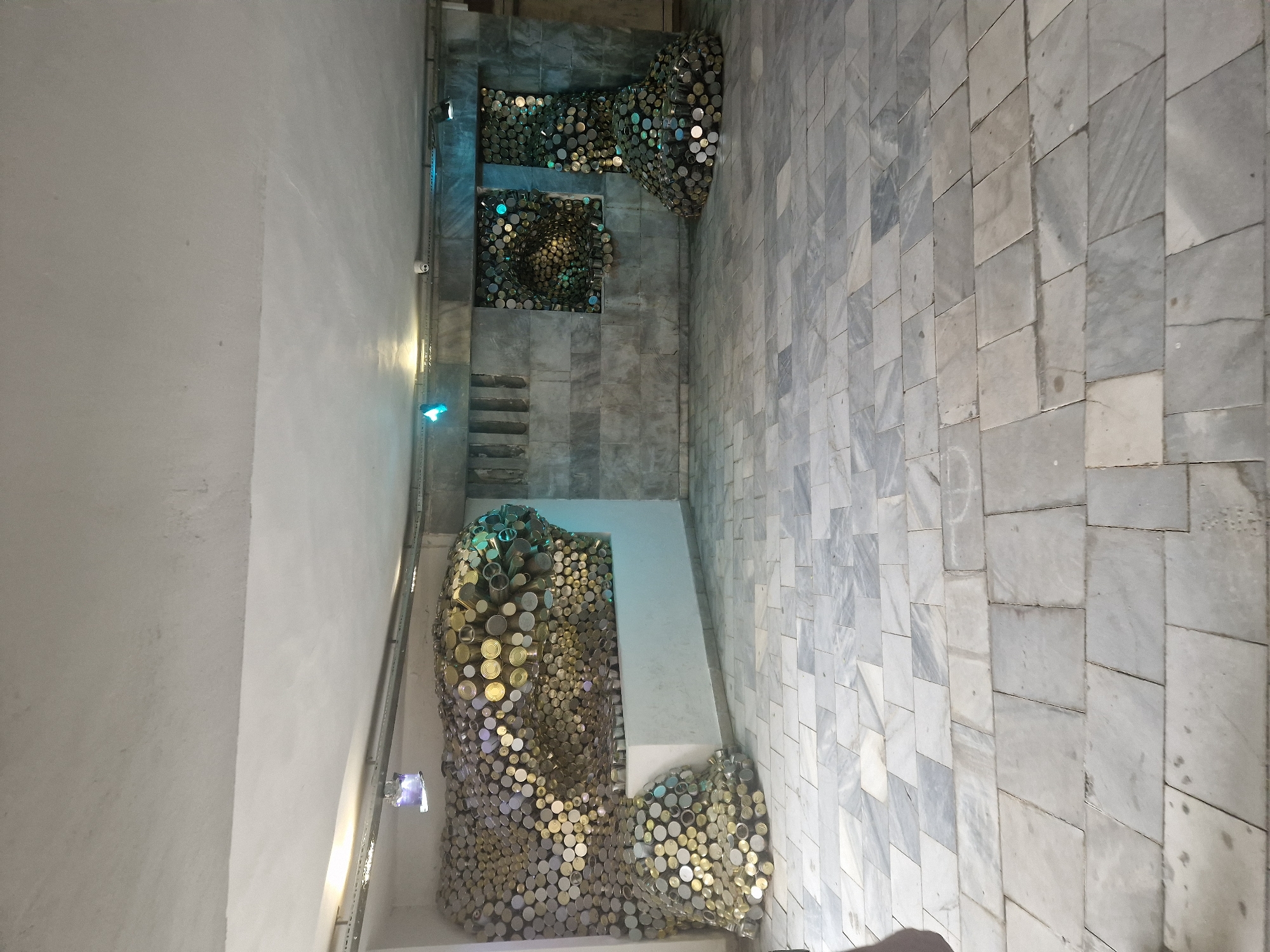 This image shows an indoor space with a gray marble tile floor and light gray walls. A sculpture composed of many round, metallic objects, possibly cans or lids, is on the left side of the image. The objects are arranged in groupings at various points on the wall. There are also metal rails along the wall. There are two additional groupings of the metallic round objects higher up the wall closer to the back of the image. A small staircase can also be seen leading up to the two metal sculptures. The location is Unknown, Unknown.