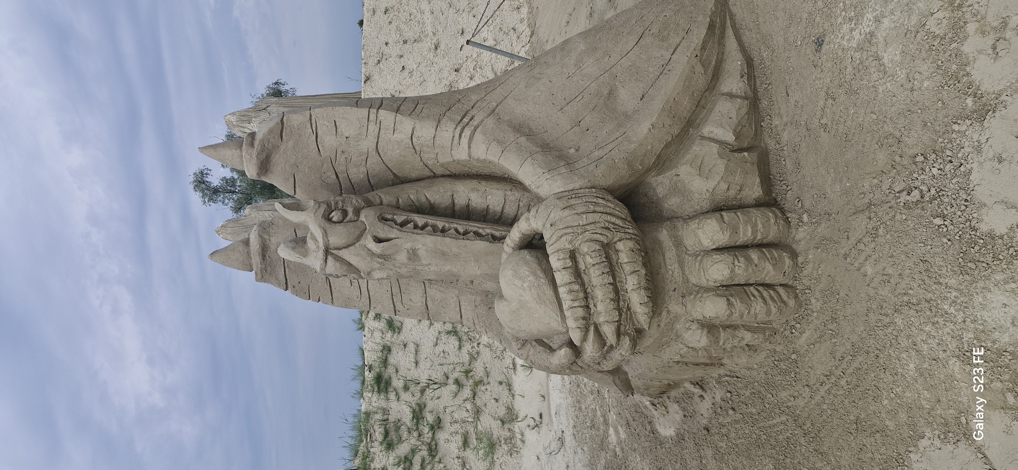 This image shows a sand sculpture of a dragon holding a heart. The sculpture is located on a sandy beach, with a cloudy sky overhead. The dragon has sharp teeth and scales, and its wings are spread wide. The heart is made of sand and is being held in the dragon's claws. The sand sculpture is a detailed representation of a mythical creature, and it is surrounded by a natural environment. The text "Galaxy S23 FE" is visible in the bottom left corner of the image, indicating that the image was taken with a Samsung Galaxy S23 FE phone.