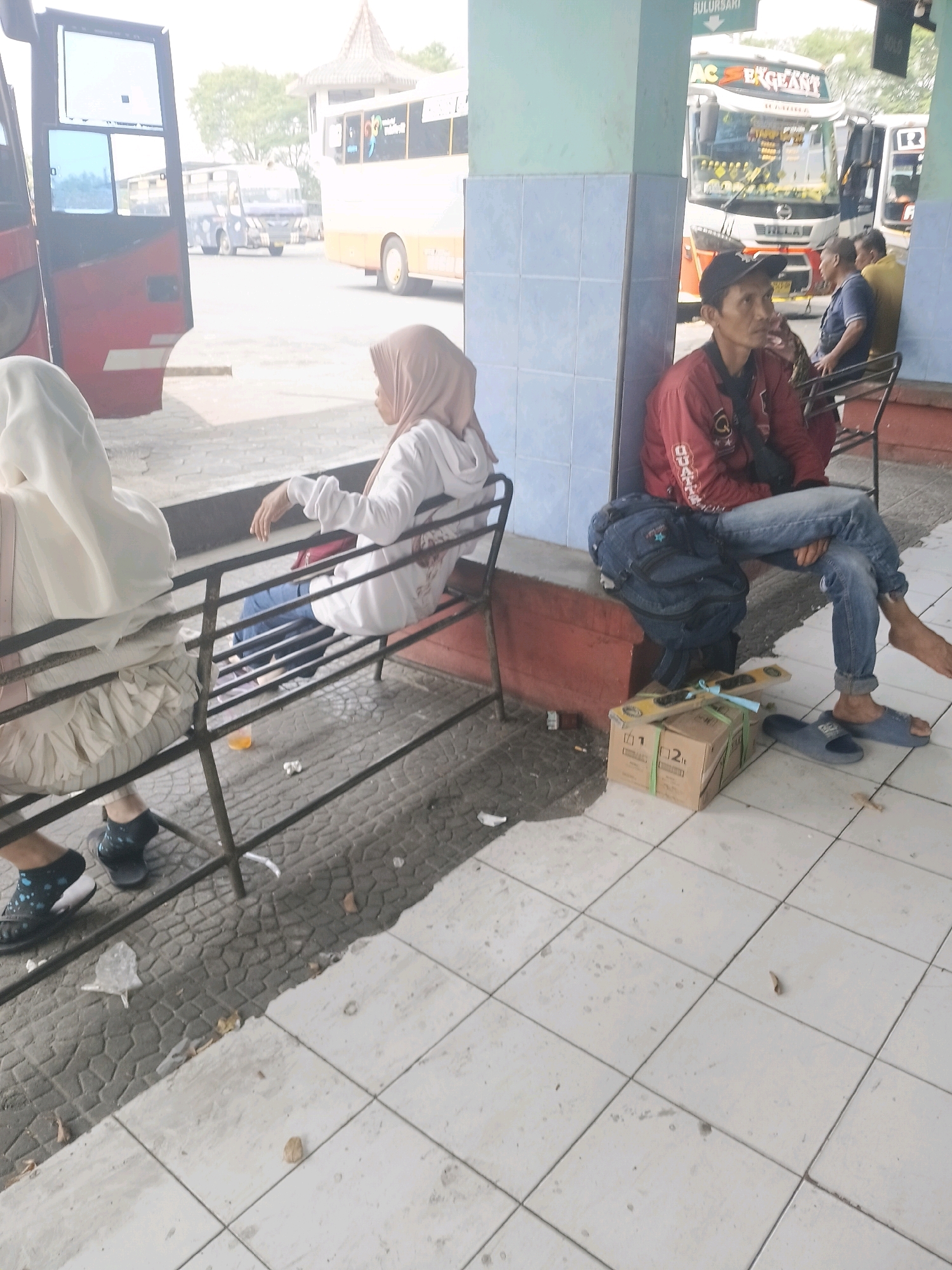 The image depicts a bus station in Kuripan, Indonesia. In the foreground, a man wearing a red jacket and blue jeans sits on a bench, looking to his right. A dark backpack is placed behind him, and a cardboard box with green strapping rests on the ground in front of him, next to a pair of blue slippers. To the left, a woman in a white hoodie and blue jeans sits on the same bench, facing away from the camera. Another woman, partially visible on the far left, wears a white hijab and a cream-colored dress, also sitting on the bench.
In the background, several buses are parked. One bus, partially visible on the far left, is red and white with its door open. Further back, a white and orange bus with the text "AC ELEGANT" is prominent, with other buses in the vicinity. The environment appears to be an outdoor waiting area, with a tiled floor and a covered structure above. The weather seems to be overcast or hazy.