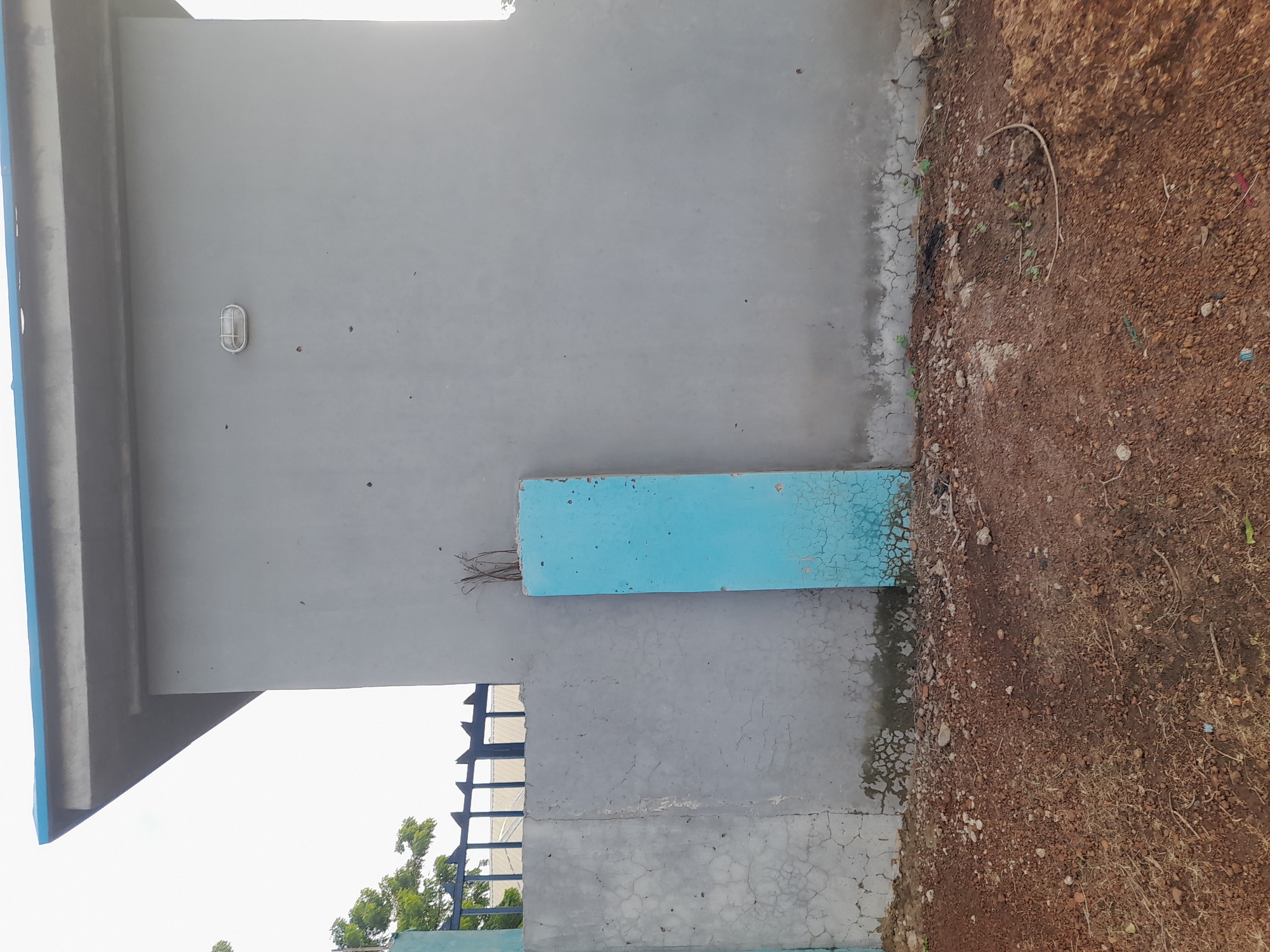 The image shows a light gray building with a blue trim along the roofline. An oval-shaped light fixture is mounted on the side of the building. In front of the building is a gray concrete wall, partially covered by a blue rectangular pillar that appears to be a decorative element or a remnant of a past structure. The ground in front of the wall and pillar is covered in reddish-brown soil, gravel, and some dry vegetation. A black metal fence is visible behind the gray wall and to the left of the pillar. The sky is overcast. The scene appears to be an outdoor area in Ibi, Nigeria, possibly a compound or a public space.