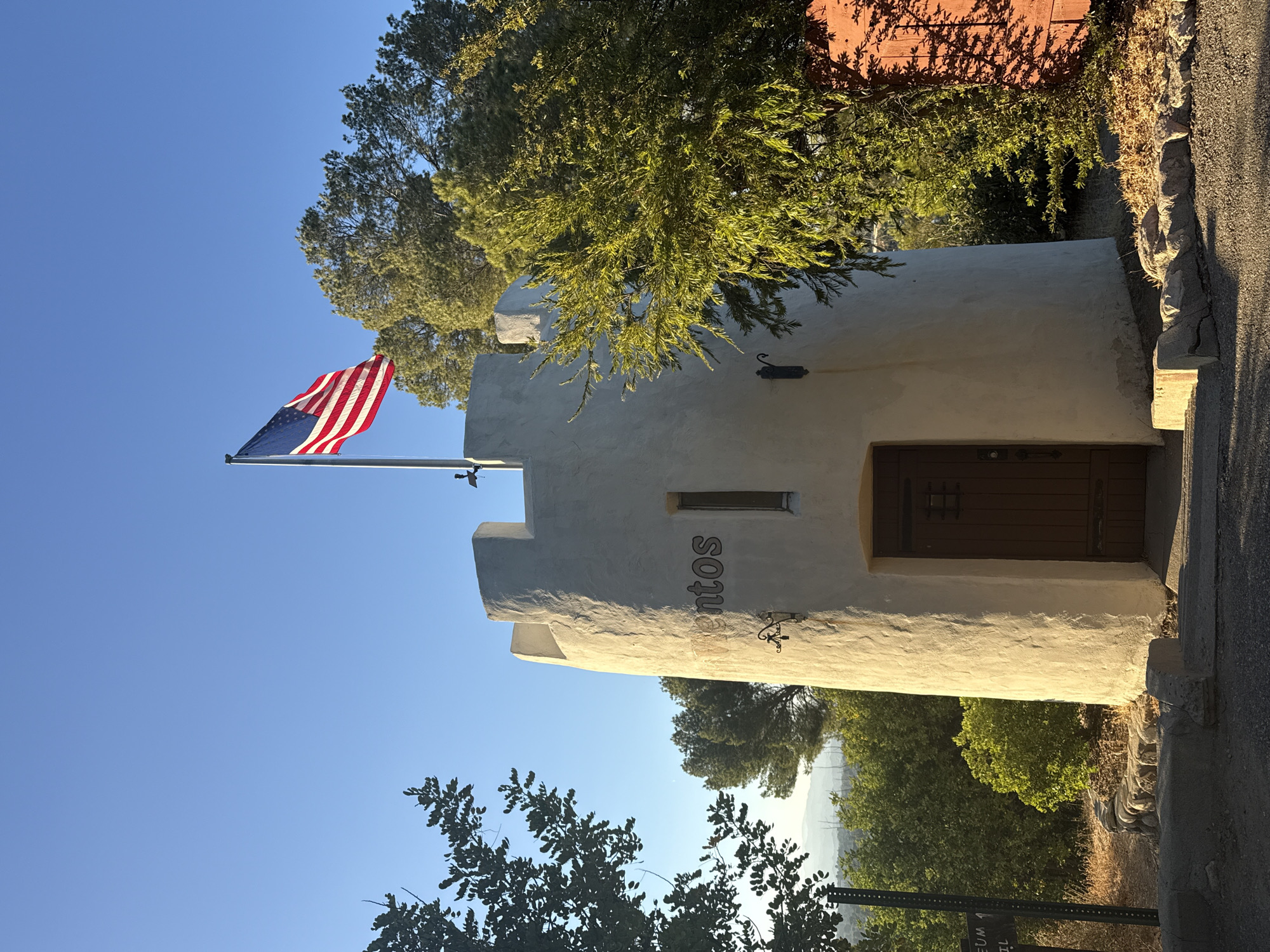 The image depicts a historic stone tower, likely part of a museum, in Santa Clarita, California, United States of America. The tower is white with a wooden door and a small window. An American flag flies from a pole on top of the tower, silhouetted against a clear blue sky. The tower is surrounded by trees and foliage, with hills visible in the background. A signpost, partially obscured by trees, is visible to the left of the tower. The lighting suggests it is early morning or late afternoon, with the sun casting long shadows.
