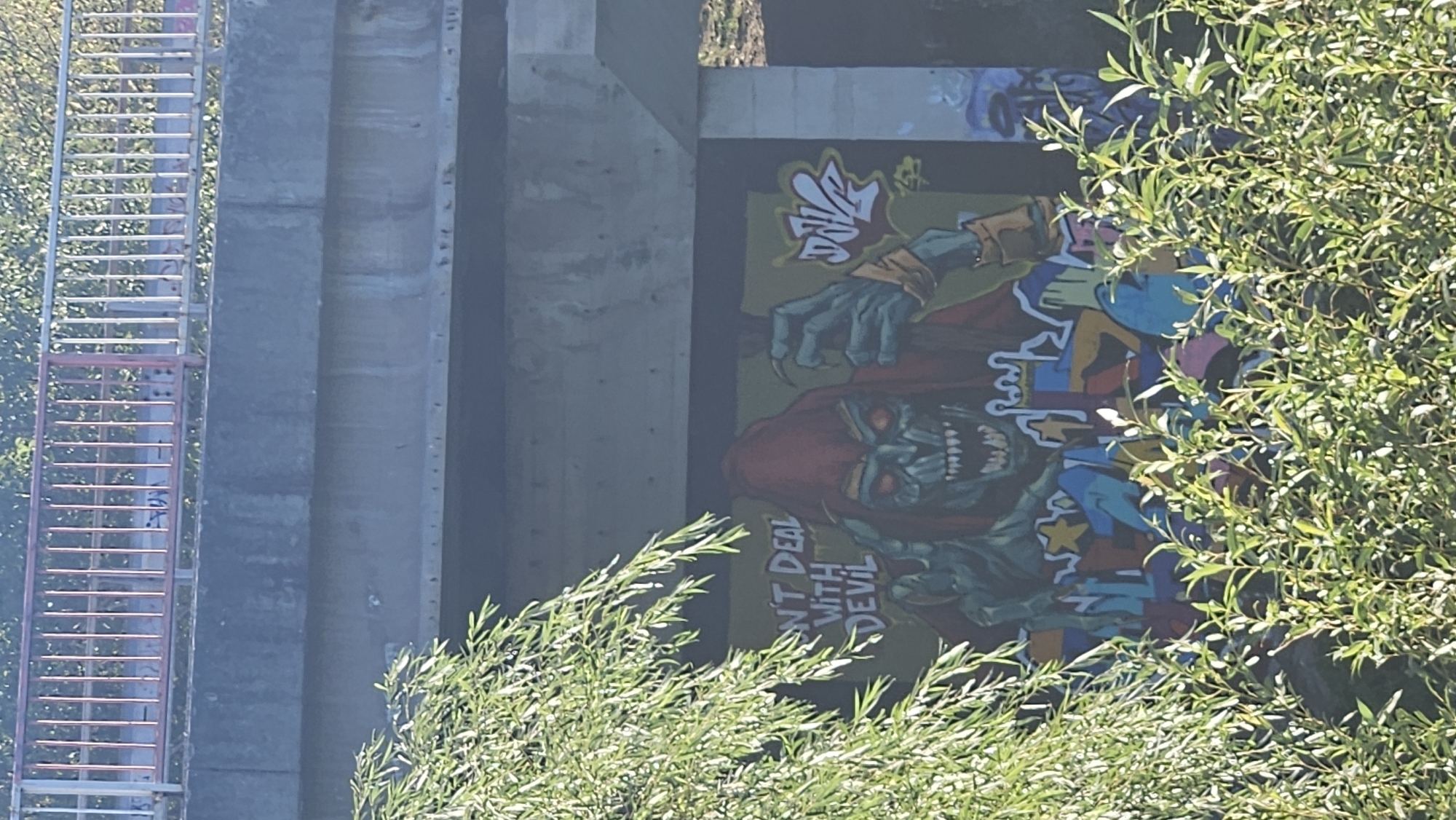 The image shows a graffiti mural of a green-skinned, monstrous figure wearing a red hood, with sharp teeth and glowing red eyes. The figure's hands are clawed, and it appears to be holding an object. Text above the figure reads "DON'T DEAL WITH DEVIL". The mural is painted on the side of a concrete structure, possibly a bridge underpass. The lower part of the mural is obscured by green foliage. The scene is likely located in Prešov, Slovakia, given the context.