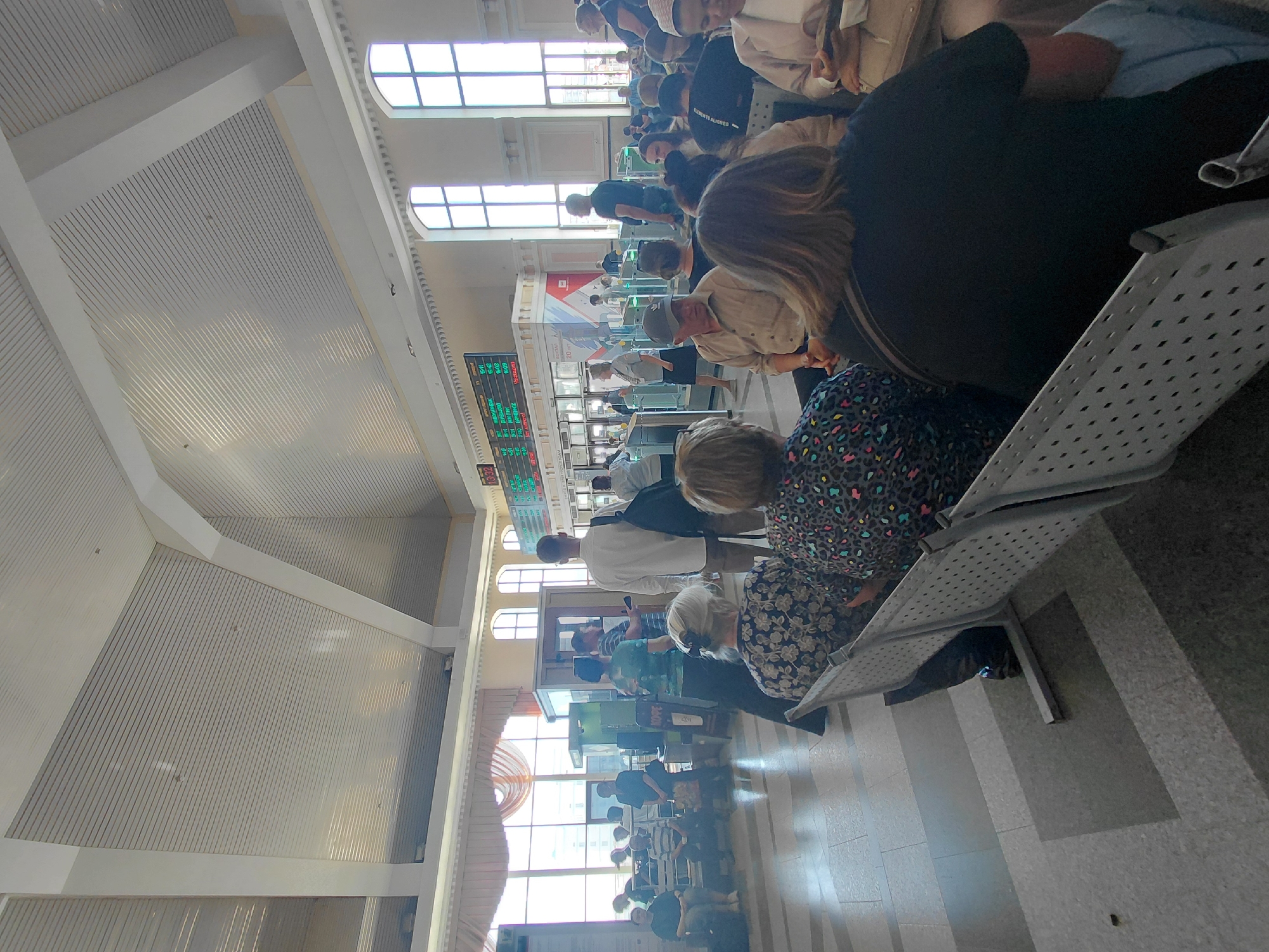 A group of people are seated and standing in a large, open indoor space, likely a train station or airport terminal. The architecture features a high, vaulted ceiling with white paneling and exposed beams. Large arched windows line the walls, letting in natural light. A digital display board showing departure times and destinations is visible in the background. People are dressed casually, and some are looking at their phones or interacting with each other. The overall atmosphere is one of waiting and transit.