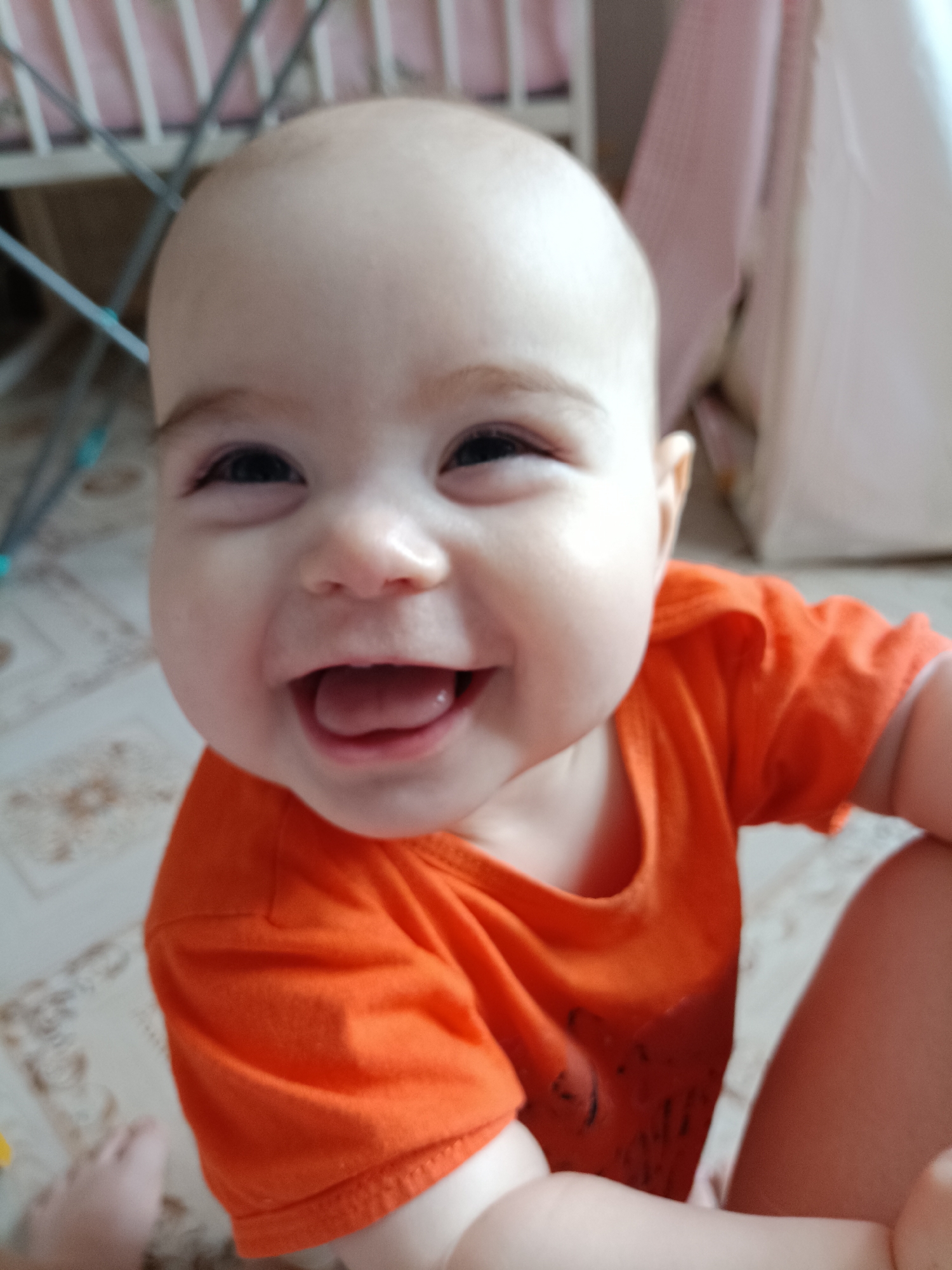 A close-up shot shows a baby with a bald head, dark eyes, and a wide, toothless smile, revealing their tongue. The baby is wearing an orange t-shirt and appears to be held or sitting in someone's lap, as part of an adult leg is visible in the lower right corner. The background includes parts of a white crib with a pink lining and a patterned floor. The location cannot be determined from this image.