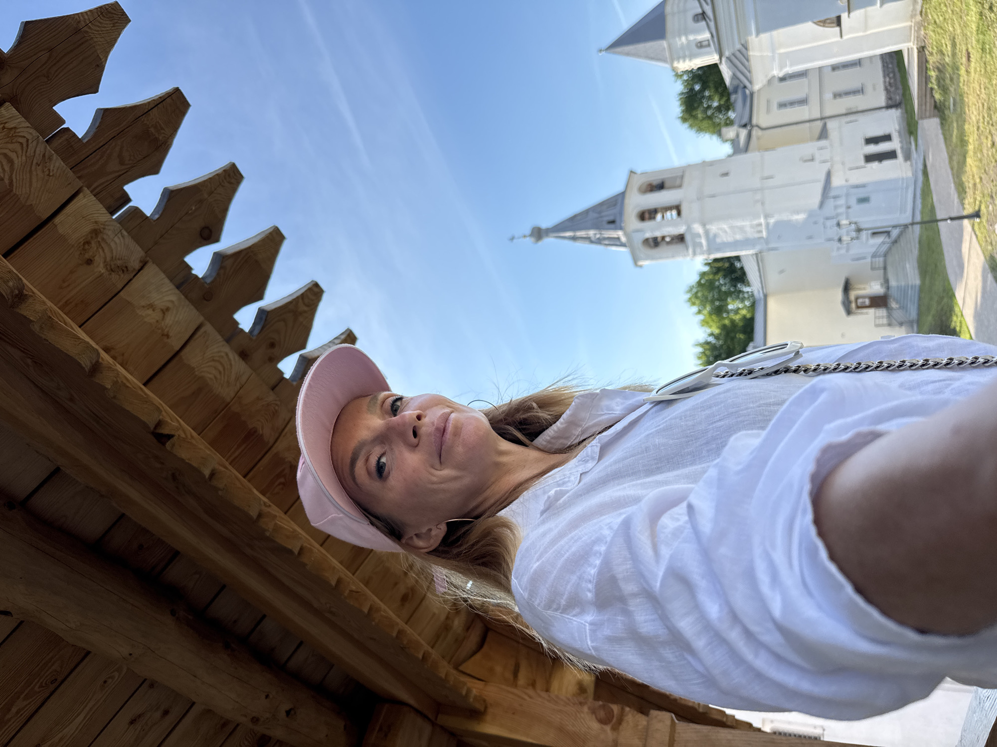 A woman in a pink baseball cap and white shirt is taking a selfie from under a wooden overhang. She has blonde hair and is wearing hoop earrings. In the background, a white bell tower with a grey spire is visible, along with other white buildings with grey roofs. Green trees are also present in the background, and the sky is blue. The architecture suggests the location is the Novgorod Kremlin in Veliky Novgorod, Russia. The perspective of the selfie is low-angle, looking up at the woman and the wooden structure.