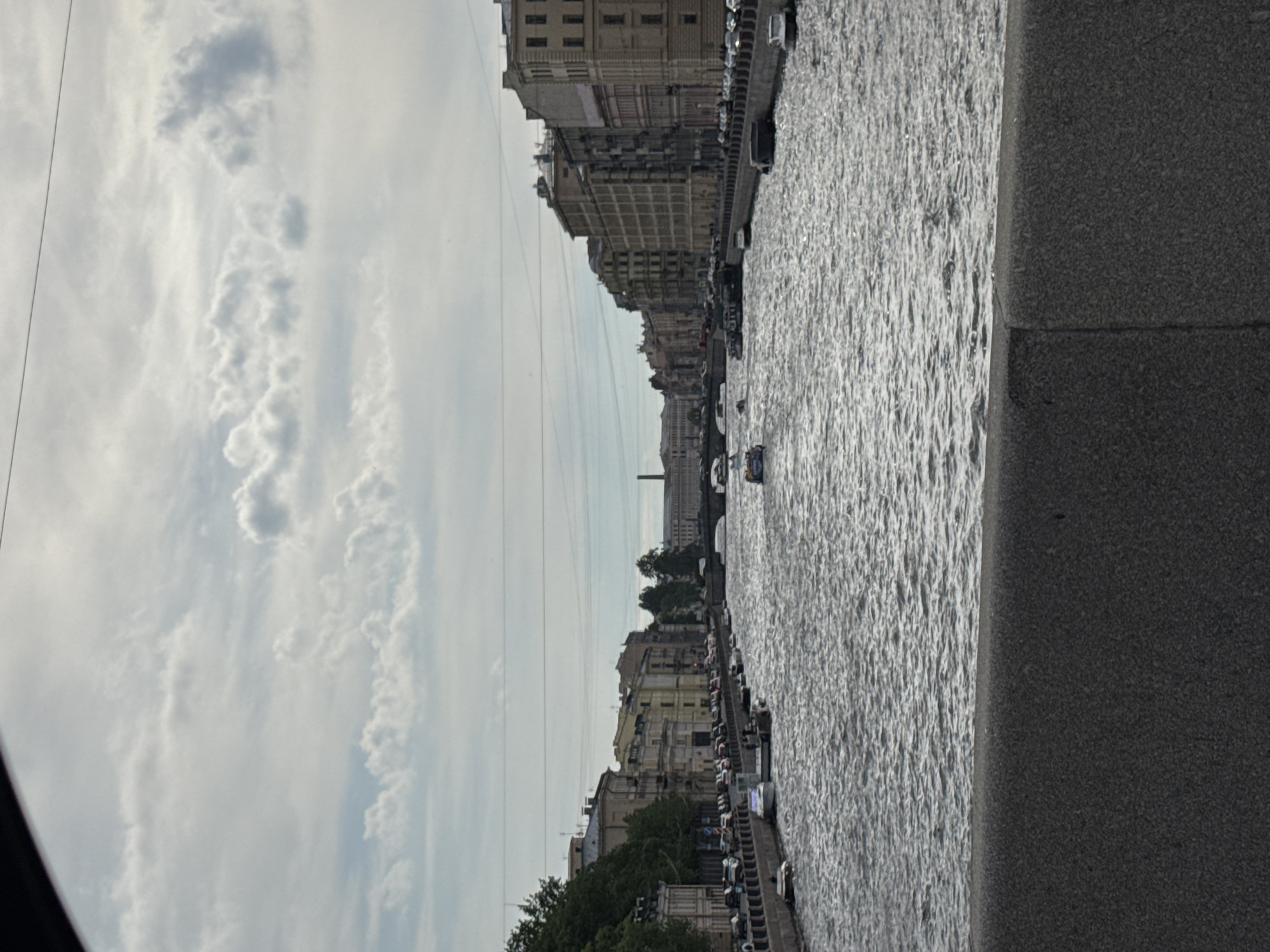 This image presents a wide view of a cityscape along a river or canal in Saint Petersburg, Russia, observed from a stone embankment. The perspective looks down the waterway, with architectural buildings lining the far bank under a cloudy sky.
The upper portion of the frame is dominated by a bright, overcast sky filled with various cloud formations, from wispy streaks to denser clusters. Several thin, parallel lines, likely power or communication wires, traverse the sky horizontally.
The body of water, prominent in the middle and right-hand side, appears rippled and textured, reflecting the diffuse light from the sky. A small, light-colored boat is visible on the water in the middle distance, moving upstream and leaving a subtle wake.
Along the distant bank, a continuous row of multi-story buildings features classic European architectural styles common to Saint Petersburg, with many windows and decorative elements. These structures appear to be a mix of residential and commercial establishments. A line of parked vehicles is discernible along the embankment in front of these buildings, and some trees and greenery are interspersed among them. In the far distance, a faint silhouette of a large, possibly domed or columned building can be seen on the horizon, hinting at a major city landmark.
The immediate foreground on the right consists of a dark, textured stone embankment, likely granite. The lighting suggests it is daytime, under an overcast but bright sky. No individuals or visible text are clearly identifiable in the scene. The image captures a serene urban landscape during what appears to be a cloudy day.