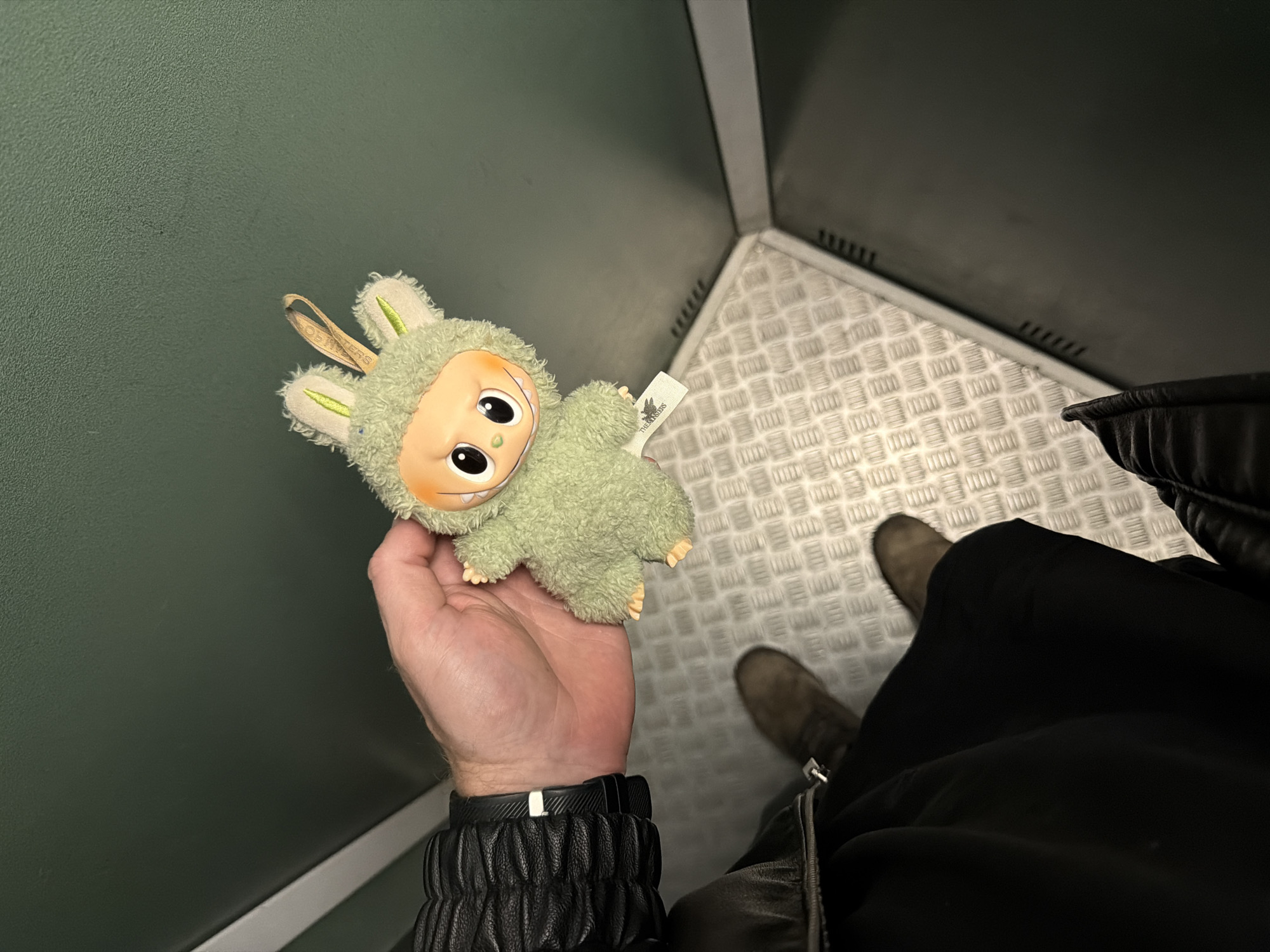 The image shows a person standing inside a small elevator or service lift. The interior walls are a dark green color, and the floor is made of a textured metal plate. The person is holding a small, green plush toy with a light-colored face and large eyes in their left hand. The person is wearing a black leather jacket, black pants, and grey boots. A smart watch is visible on their left wrist. The image was taken at an angle from above, capturing the person's hand, arm, and part of their lower body, as well as the plush toy and the surrounding elevator interior. There is no specific information in the image to indicate that the photo was taken in Saint Petersburg, Russia.