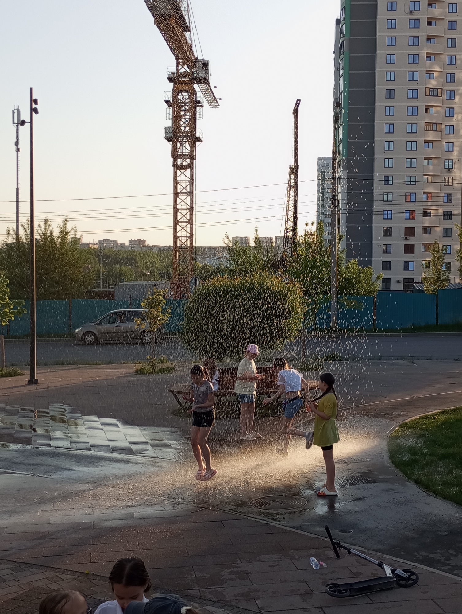 The image shows a group of children playing in a water fountain in what appears to be a public area. There are at least five children, and they are standing and moving in the spray of the water. The area is paved, and there is a scooter and a water bottle lying on the ground nearby. In the background, there are several tall buildings, two construction cranes, trees and bushes. A car is parked on the road behind the fence. The photograph was taken in the city of Pervomaysky, Russia.