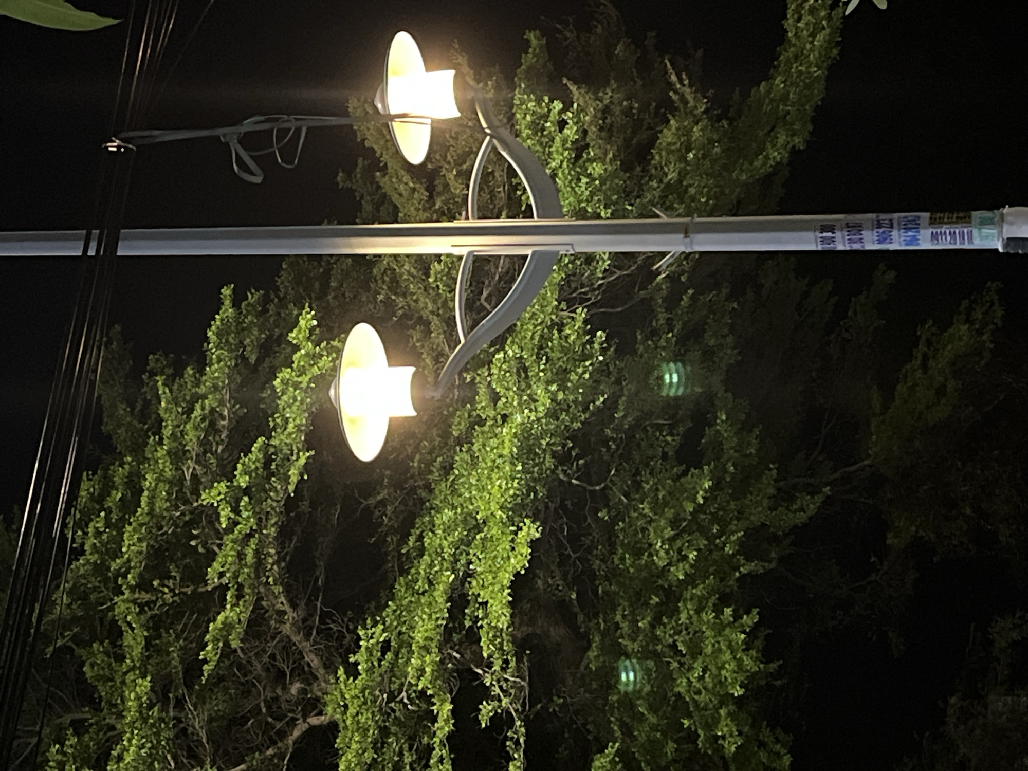 In Phu Thuy Ward, Vietnam, a street lamp stands tall against a dark night sky, illuminating a dense, leafy tree. The lamp features two distinct light fixtures, each with a wide, flat shade casting a warm, yellow glow. One light is positioned higher and to the right, while the other is slightly lower and to the left, attached to the same pole. The pole itself is a slender, metallic structure, with a series of stickers or labels adhered to its lower section. These labels are predominantly blue and white, displaying what appear to be alphanumeric codes or numbers. The tree, abundant with vibrant green leaves, forms a thick canopy behind the lamp, its branches reaching outwards. The overall scene is one of urban nighttime illumination, where artificial light interacts with natural foliage.