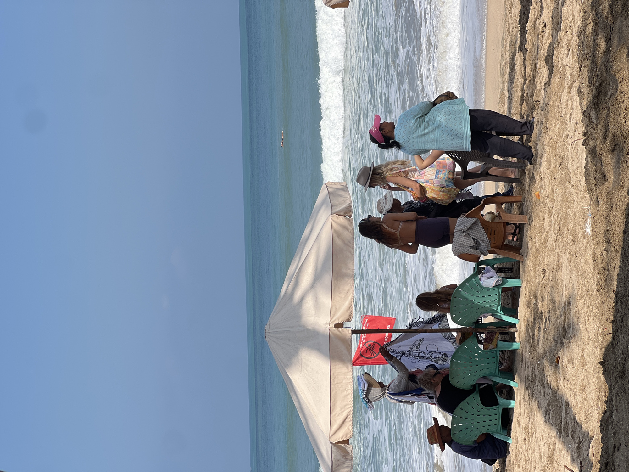 In Kuta, Indonesia, a group of people are gathered on a sandy beach near the ocean under a large umbrella. The ocean waves are breaking on the shore. Several individuals are standing and sitting, some appearing to be vendors selling merchandise, such as cloths and hats. There are also people who appear to be tourists, enjoying the beach atmosphere. One person is seen swimming in the ocean in the distance. The scene depicts a typical day at a popular Indonesian beach.