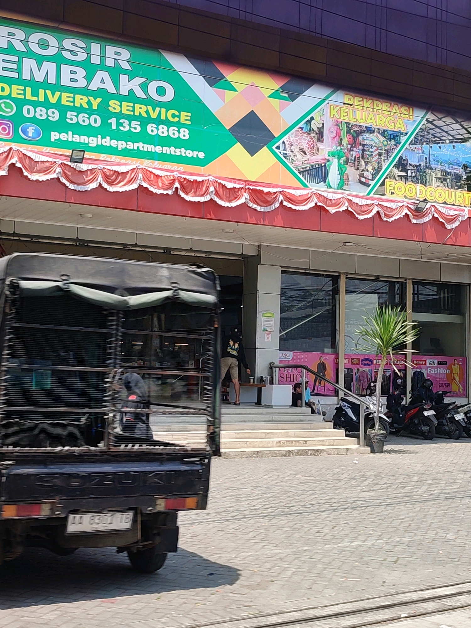 The image is taken from a street-level perspective, showing the facade of a building in what appears to be a commercial area. A black Suzuki pickup truck with a caged rear is prominently featured in the foreground, moving from left to right. The truck has a license plate that reads "AA 8301 TB".
Above the truck, the building displays a large, green and yellow banner with text in Indonesian. The text includes "ROSI" and "EMBAKO", followed by "DELIVERY SERVICE" and a phone number "089 560 135 6868". Below this, social media icons are visible, including an Instagram logo and a Facebook logo, alongside the text "pelangidepartmentstore". Further down, a decorative red and white valance runs across the top of the storefront.
The storefront itself has large glass windows, through which some interior displays are visible. Posters advertising clothing and fashion are displayed on the windows, with text such as "Fashion" and "SHIO". Several motorcycles are parked in front of the building, and a few people are seen standing and sitting near the entrance. A small potted plant is located to the right of the entrance. The ground is paved with patterned tiles. The background shows the upper facade of a modern building with dark windows, suggesting a multi-story structure. The lighting indicates it is daytime, with bright sunlight casting shadows.
While the specific location of Tempurejo, Indonesia is not explicitly visible in the image itself, it is inferred from the context provided. The architectural style and signage suggest a Southeast Asian setting.