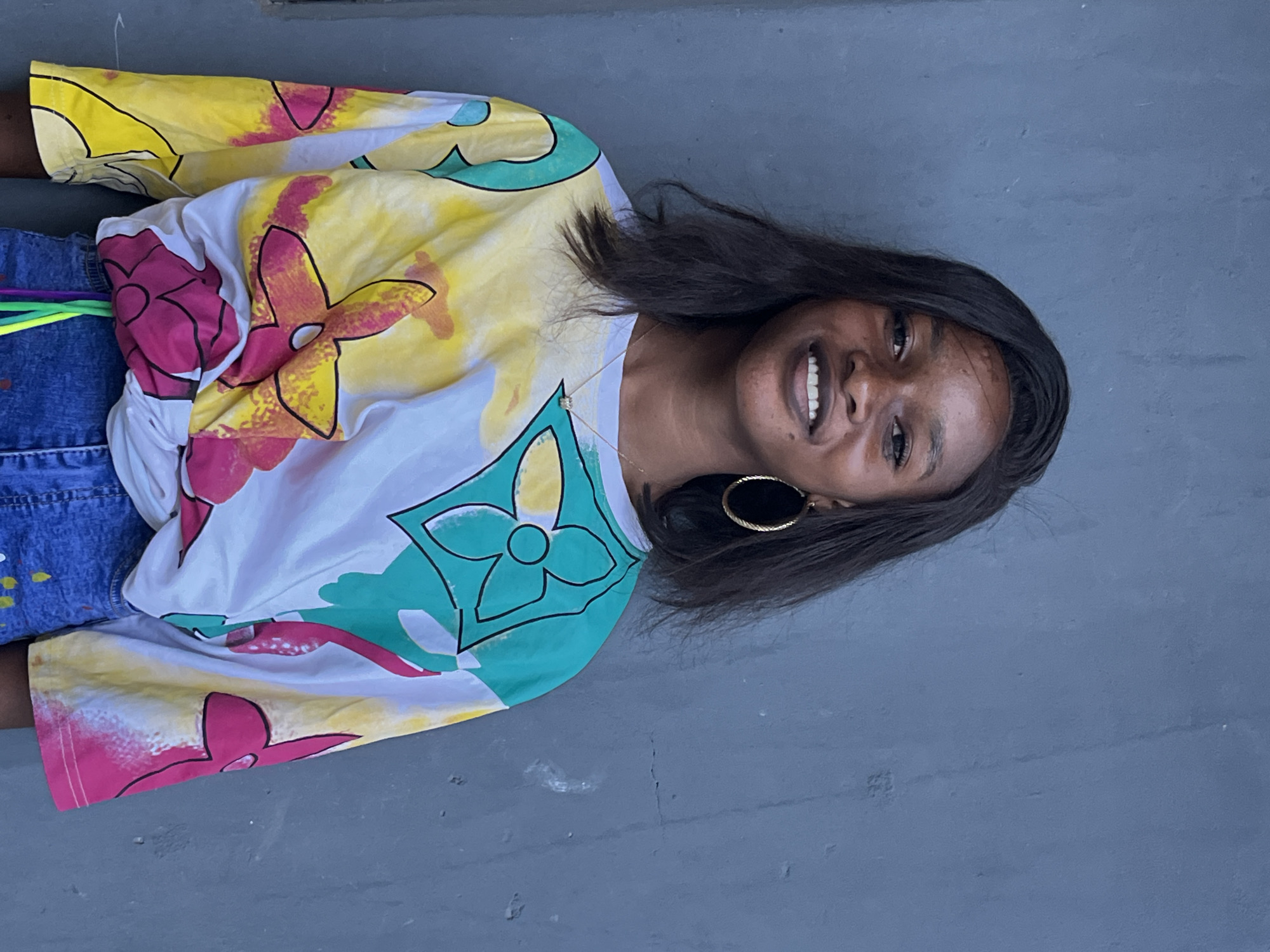 The image is an eye-level portrait of a woman, angled slightly to the right, against a grey textured wall. She is wearing a short-sleeved, colorful shirt with floral designs in shades of yellow, pink, and teal. The woman has dark hair, hoop earrings, and a necklace. She is smiling, revealing her teeth. Part of her blue jeans is visible on the left side of the image, with colorful accents on the fabric. The background is a plain, grey wall, suggesting an indoor setting, possibly in a residence or building in Badagry, Nigeria.
