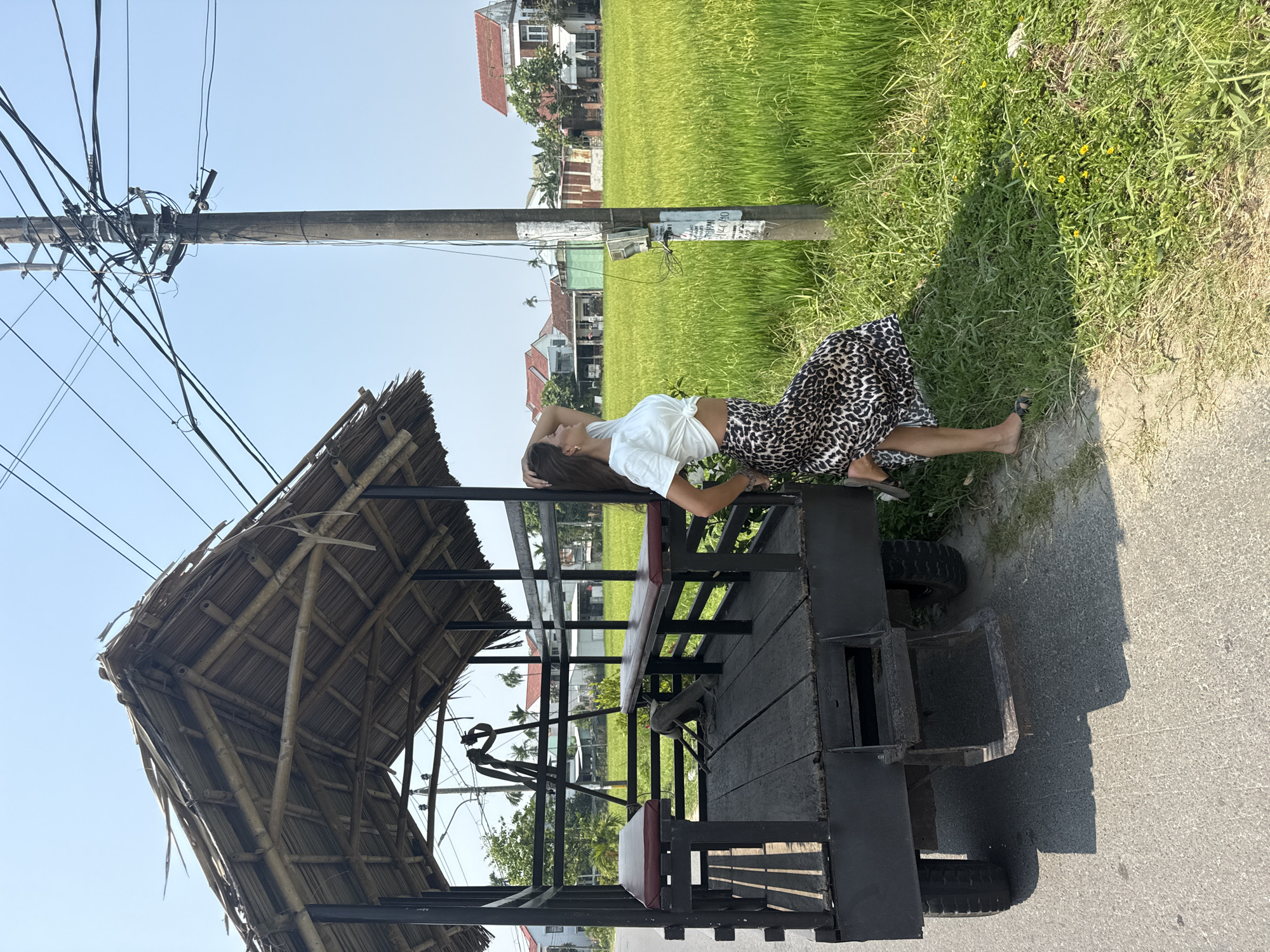 A woman reclines on the back of a rustic, canopied vehicle on a paved road in a rural area outside of Da Nang, Vietnam. The vehicle features a bamboo canopy and is parked near a grassy area and a utility pole with power lines. In the background, there is a lush green rice field and a few houses. The woman is wearing a white top and a leopard-print skirt.