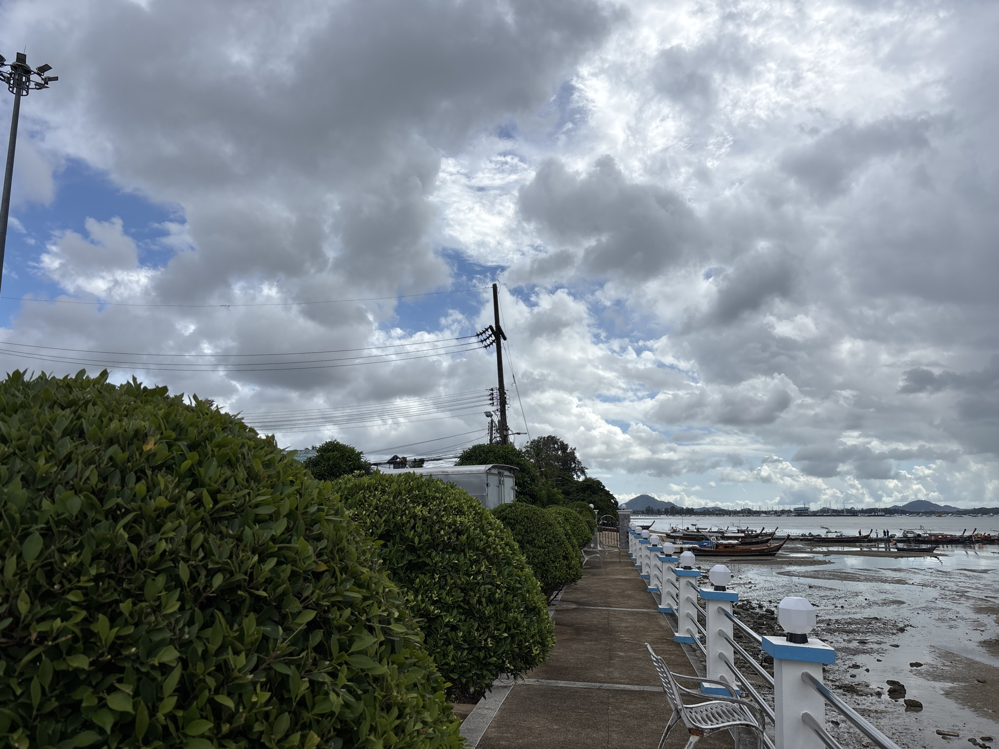 The image depicts a waterfront promenade in Rawai, Thailand, on a cloudy day. A paved walkway runs along the left side of the frame, bordered by manicured green bushes. On the right side, a white and blue railing separates the walkway from the beach area. The beach is exposed due to low tide, revealing wet sand, rocks, and shallow water. Numerous long-tail boats are moored in the shallow water, some closer to the shore and others further out. In the background, a distant coastline with hills can be seen under the overcast sky, which is filled with large, dramatic clouds. A few utility poles with overhead wires are visible along the edge of the promenade, and a metallic trailer is parked behind the bushes. There are no people clearly visible in the immediate foreground or midground of the image, though some figures can be faintly discerned near the boats in the distance.