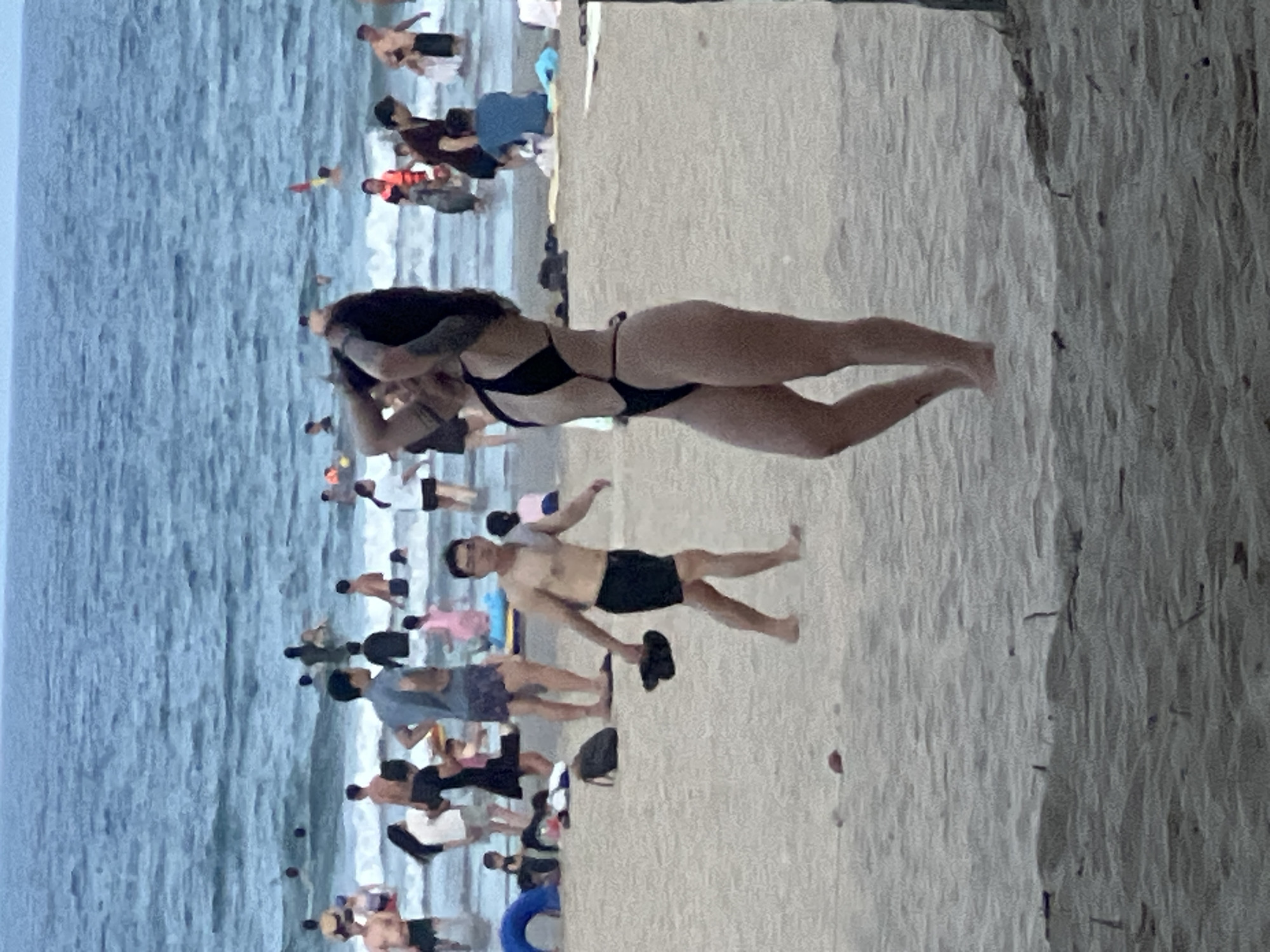 The media file captures a lively beach scene in Đà Nẵng, Vietnam. The image is oriented vertically. On the left side, a vast expanse of the ocean with gentle waves suggests a pleasant day for swimming or wading. The water appears a muted blue-gray, indicating possibly overcast conditions or early morning/late afternoon light. Scattered throughout the water and the shoreline are numerous people enjoying the beach.
The foreground and right side of the image focus on the sandy beach. Prominently featured is a woman in a black bikini, her back to the viewer, her long dark hair cascading down. She appears to be posing or adjusting her swimwear. Further up the beach, a man in black swim shorts is visible, holding a pair of flip-flops in his hand. He is walking across the sand.
The beach is populated with many individuals and small groups, some wading in the water, others walking along the shore or simply relaxing on the sand. The attire of the beachgoers, predominantly swimwear, is typical for a warm, coastal environment. The presence of what appears to be a blue flotation ring suggests recreational activities.
The overall atmosphere conveyed is one of leisure and enjoyment, with a sense of community gathering at the seaside. The time of day is not definitively clear, but the diffused light might suggest it's not the harsh midday sun. The location is clearly identified as a beach in Đà Nẵng, Vietnam, a popular tourist destination known for its scenic coastline. No visible text is present in the image.