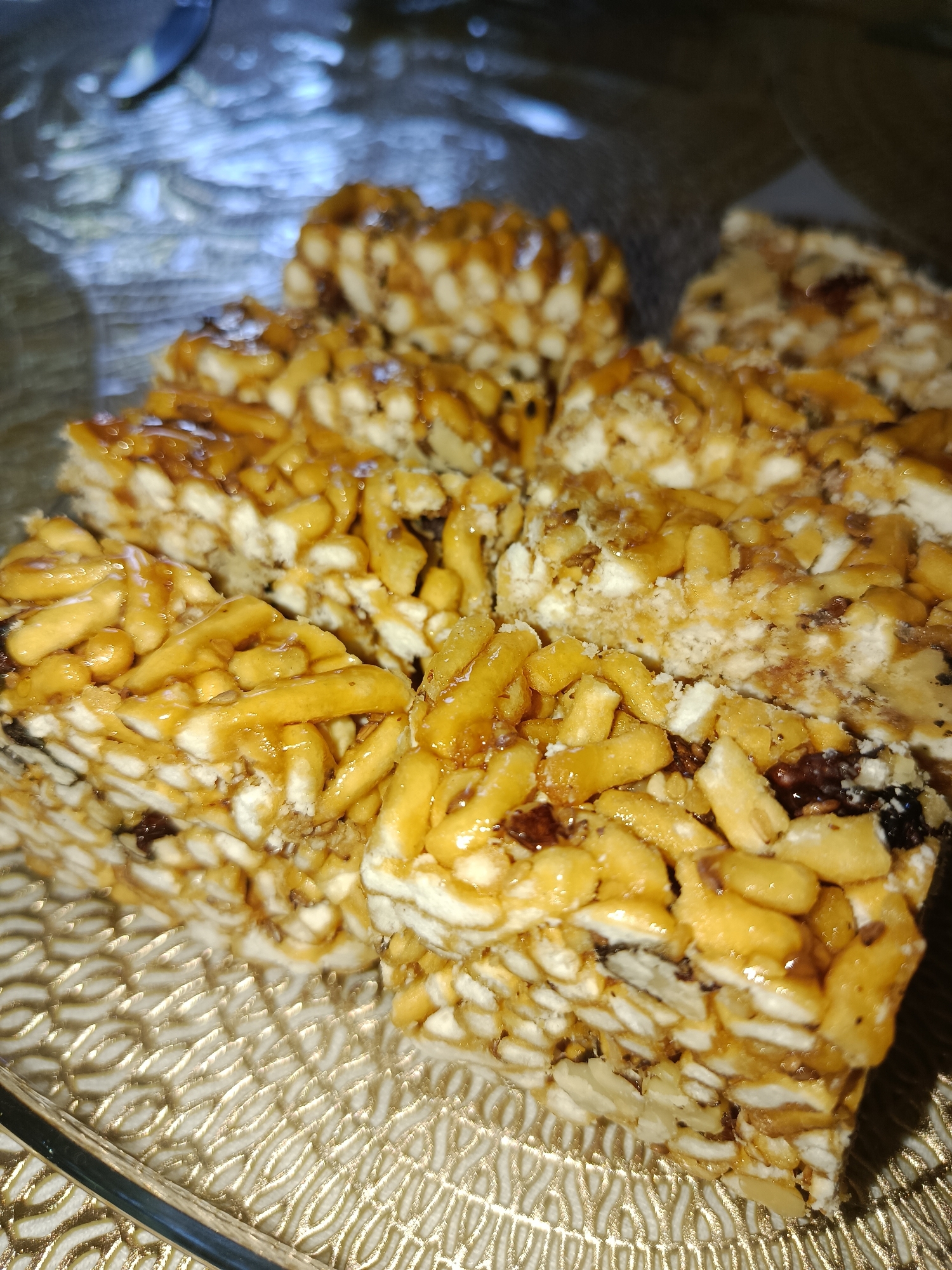 A close-up, overhead shot displays a plate of Chak-Chak, a traditional Uzbek dessert, set on a decorative glass platter. The Chak-Chak is cut into rectangular pieces, revealing layers of fried dough coated in honey or syrup. Visible ingredients include small, stick-like pieces of dough, some larger pieces resembling nuts or puffed grains, and dark specks that could be dried fruit or spices. The dessert is glossy, with a golden-brown hue. The platter has an intricate, embossed pattern that reflects the light, creating a textured surface. In the background, a blurred utensil, possibly a spoon, rests on a surface with a shimmering, light-colored texture. The overall lighting suggests an indoor setting, likely a home or a cafe in Tashkent, Uzbekistan, given the context of the dessert.