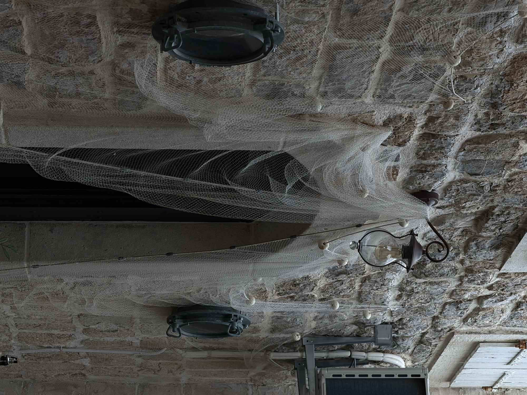 The image shows a ceiling decorated with a fishing net and nautical-themed lighting fixtures. The ceiling is constructed of rough-hewn stones, likely within an older building. Two round, metal fixtures resembling porthole lights are visible, along with an ornate, hanging lamp with a glass shade. Small, white spherical objects are interspersed within the fishing net. The environment suggests a restaurant or bar with a maritime theme, possibly located in the city of Budva, Montenegro, given the city's coastal location and association with nautical history.