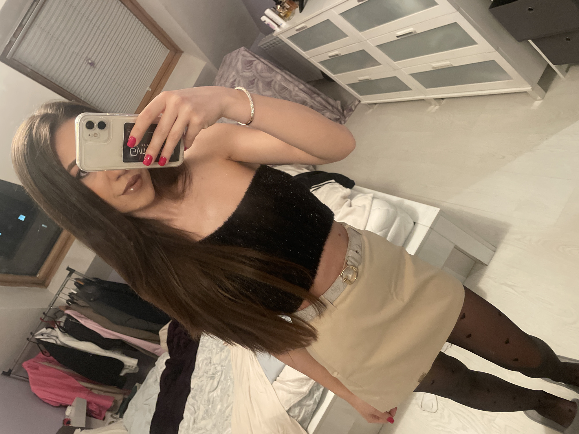 A young woman is taking a selfie indoors, likely in her bedroom. She's wearing a strapless black top, a beige skirt with a gold belt buckle, and patterned black tights. She is holding an iPhone in her right hand. The room includes a dresser, a clothes rack with various garments, and what appears to be a bed with white bedding. The environment seems domestic. The location is not discernible as being in Sofia, Bulgaria, from the visual elements of the image alone.