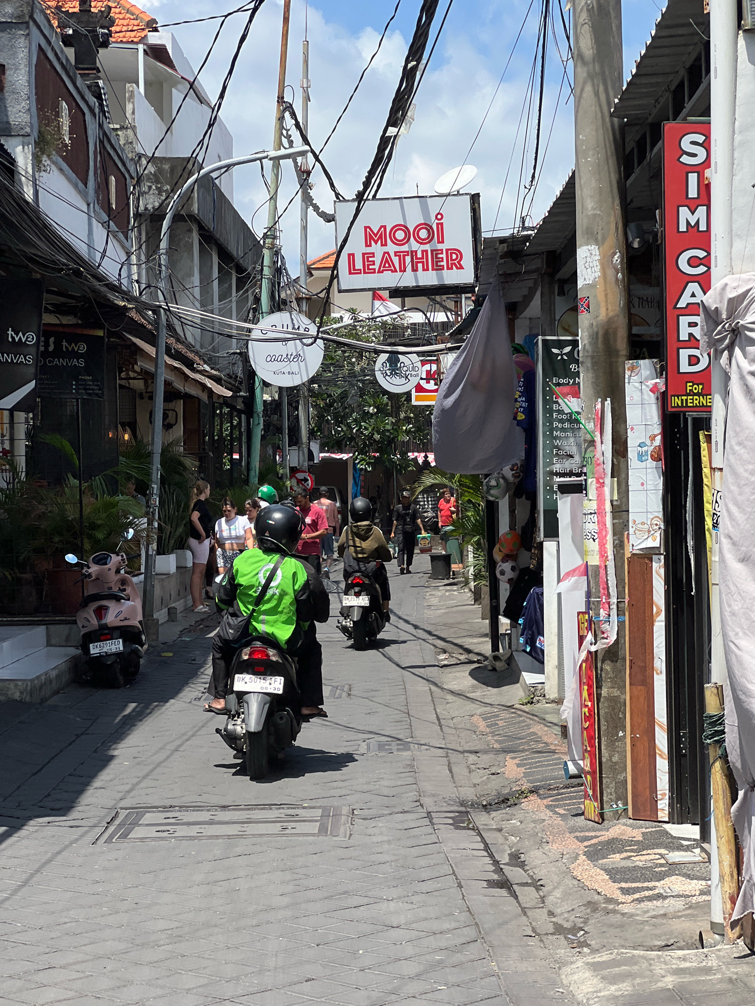The image depicts a narrow, sunlit street in Kuta, Indonesia, characterized by a dense network of overhead electrical wires. On the left side of the street, a shop displays "TWO 3 TWO CANVAS D CANVAS." Further down, a circular sign reads "CRUMP & coaster KUTA BALI." Across the street, a red sign with white lettering states "SIM CARD" and below it "For INTERNET." A person wearing a bright green jacket and a black helmet rides a motorcycle away from the camera. To their right, another person on a motorcycle is also riding away. Several other individuals are visible walking or standing along the sidewalk. Scooters are parked along the left side of the street. The street is paved with grey interlocking blocks.