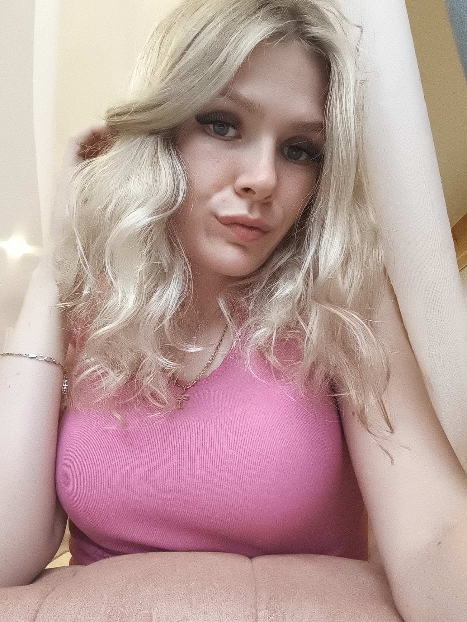 This is a selfie of a young woman with blonde, wavy hair. She is wearing a pink ribbed tank top and a necklace with a small pendant. A silver bracelet is visible on her left wrist. Her makeup includes noticeable eyeliner and mascara. She is looking towards the camera with a slightly pensive expression. The background consists of soft, diffused lighting and what appears to be light-colored fabric, possibly curtains or drapes. There are some small, out-of-focus lights in the upper left corner of the image. The setting appears to be indoors, with a warm and intimate atmosphere. The lighting suggests it could be daytime, possibly late afternoon, given the soft, ambient glow. There is no visible text in the image.