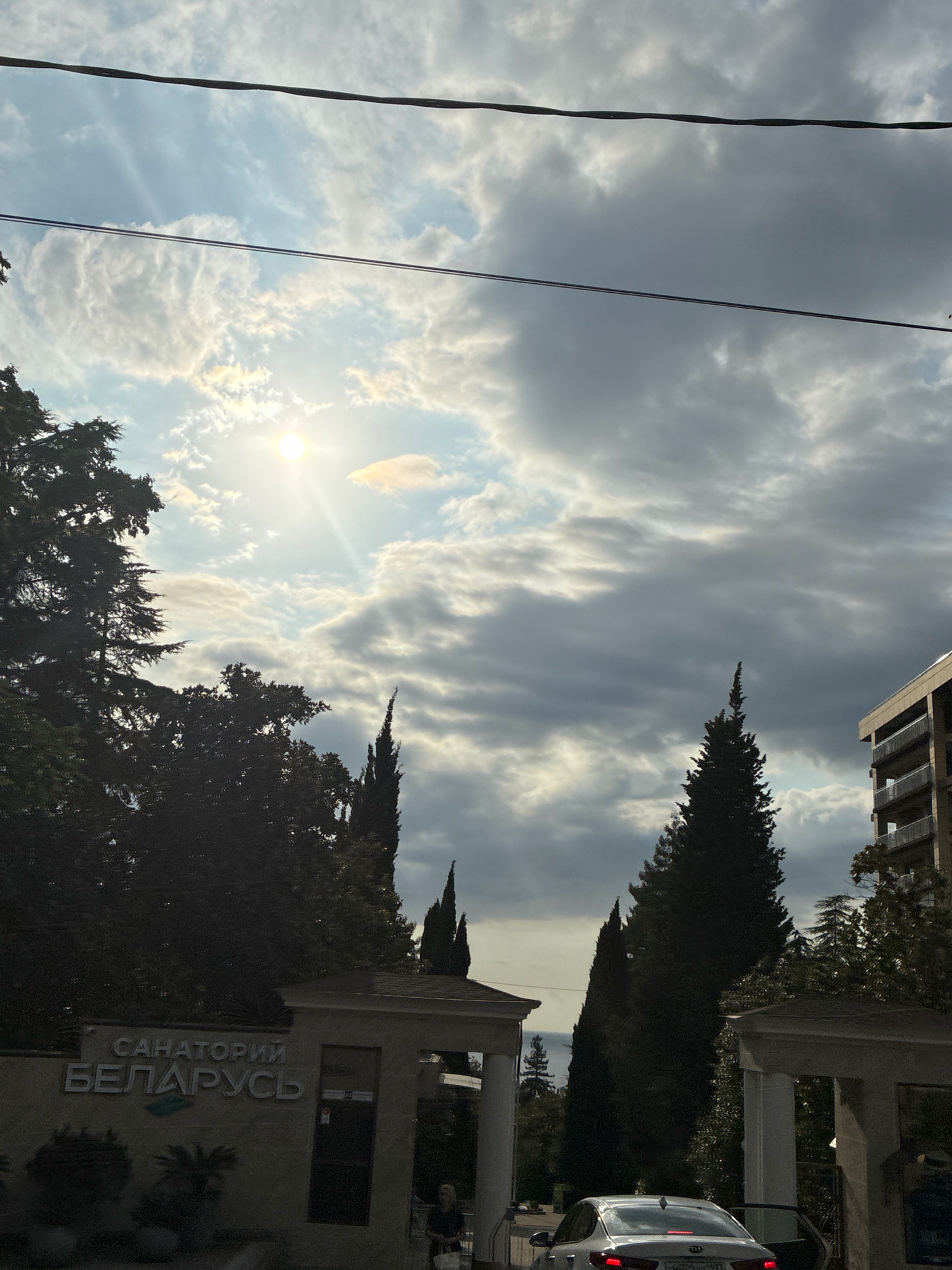 This image captures a view of the Sanatorium Belarus in Sochi, Russia. The sun is partially obscured by clouds, casting rays through the sky. In the foreground, a white car is parked with its driver's side door open. A person is standing near the car, facing away from the camera. The sanatorium's entrance gate is visible, with the name "САНАТОРИЙ БЕЛАРУСЬ" displayed on it. Tall, dark green cypress trees line the entrance and the surrounding area. In the background, a multi-story building with balconies can be seen to the right, and the sea is visible in the distance to the left. Overhead, power lines stretch across the frame.