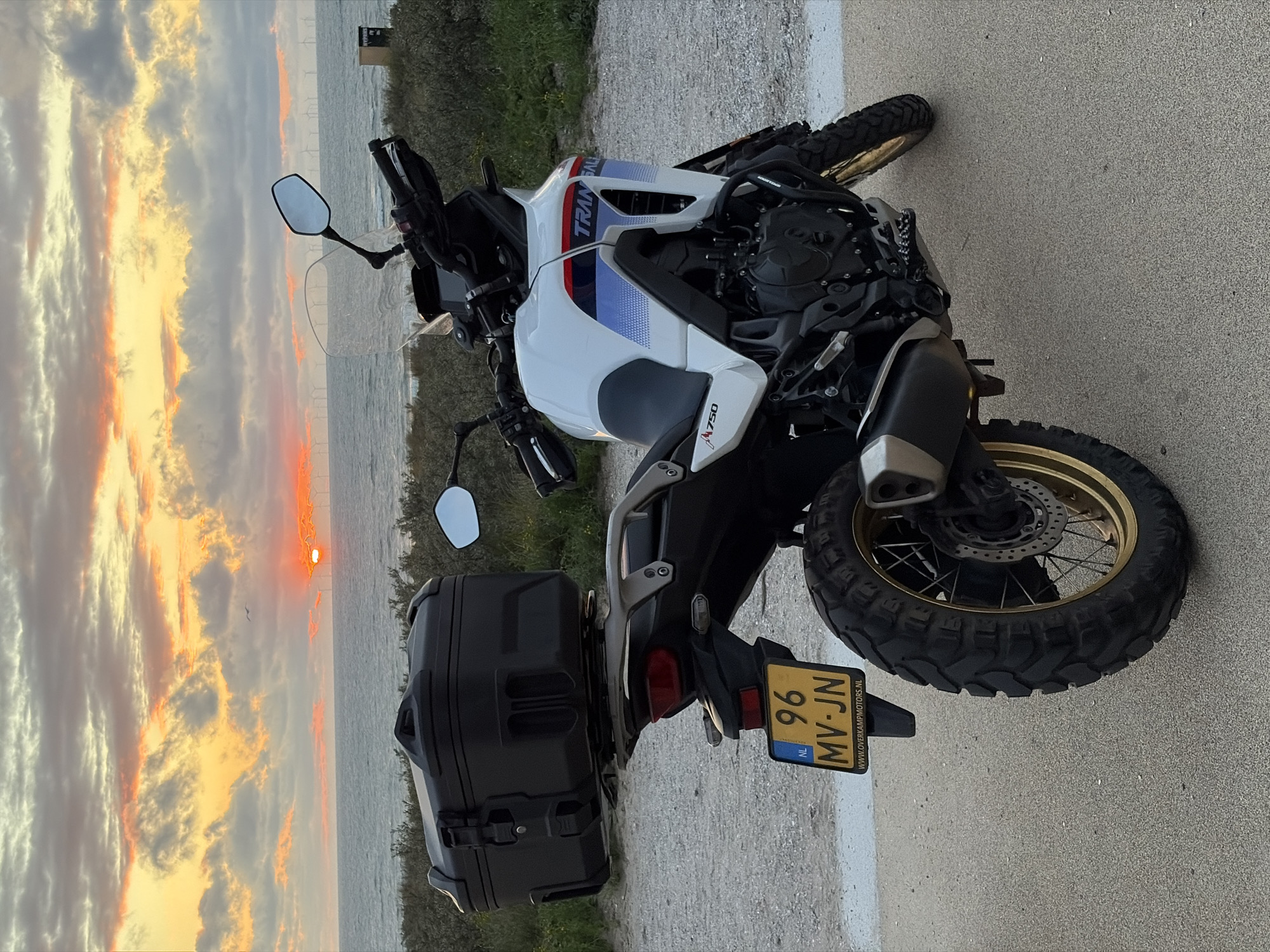 The image shows a white Honda Transalp motorcycle parked on a sandy path near a body of water, likely the North Sea. The sun is setting, casting a warm orange glow over the sky and clouds. In the distance, numerous wind turbines are visible on the horizon. The motorcycle has a black cargo box mounted on the rear, and a Dutch license plate displaying "96 MV-JN". The scene appears to be a coastal area, possibly within the municipality of The Hague, Netherlands, known for its extensive coastline and offshore wind farms. There are no people or animals visible in the image.