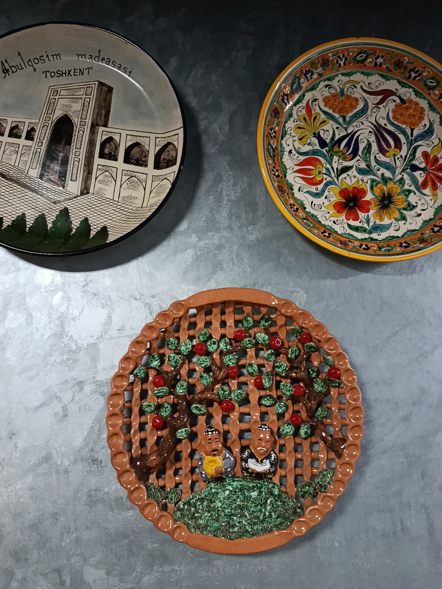 Three decorative plates are displayed on a gray surface.
The plate in the upper left features an architectural rendering of Abdulqosim Madrasasi in Tashkent, Uzbekistan, with its name written above the illustration. The plate has a black rim.
The plate in the upper right is brightly decorated with colorful floral motifs, including red, orange, and yellow flowers, set against a white background. It has a yellow rim.
The bottom plate depicts two figures sitting under trees laden with red fruit, against a background resembling a trellis. The plate's rim is brown.