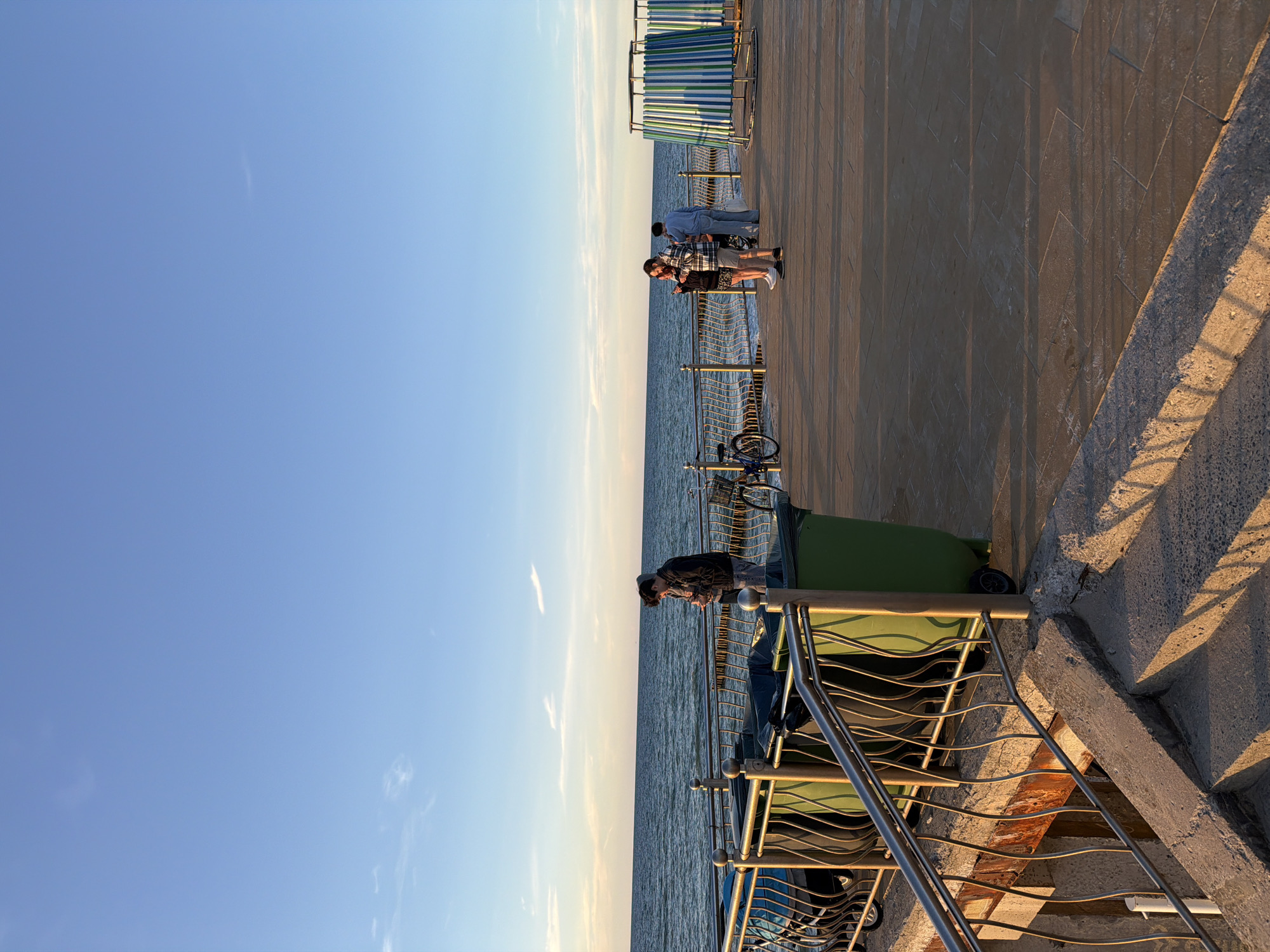 The image depicts a scene on a seaside promenade in Zelenogradsk, Russia. The foreground features a concrete staircase with a metal railing, leading up to the promenade. The promenade itself is paved with square tiles and has a metal railing along the edge overlooking the sea. Several large green trash receptacles are visible along the promenade. In the middle ground, a young person wearing a dark jacket stands near the railing, looking at their phone. Further along the promenade, a group of three people are standing together. One person is wearing a plaid shirt and shorts, another a light blue top and pants, and a third person is partially obscured. A bicycle is parked near the railing. In the background, the Baltic Sea stretches to the horizon under a clear blue sky with scattered light clouds. The sun is low, casting long shadows and illuminating the scene with warm, golden light. The image captures a moment of calm on a coastal public space.