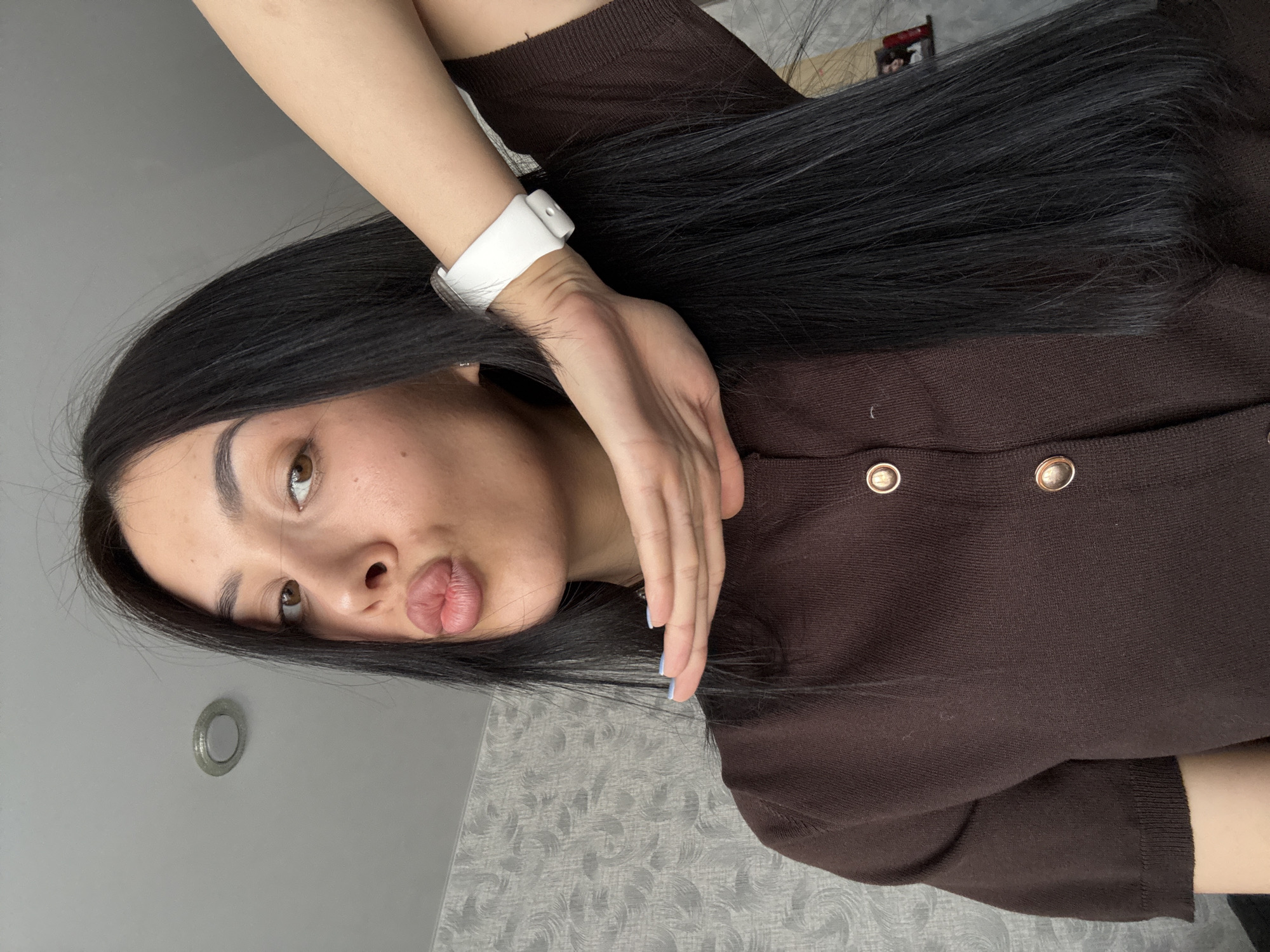 A woman with black hair and light brown eyes is posing for a selfie. She is wearing a brown button-down shirt and a white smartwatch. Her lips are puckered. The background is a light gray wall with a textured pattern. The image is taken from a slightly elevated angle. There's a light fixture visible in the lower left corner. There is no indication this image was taken in Votkinsk, Russia.