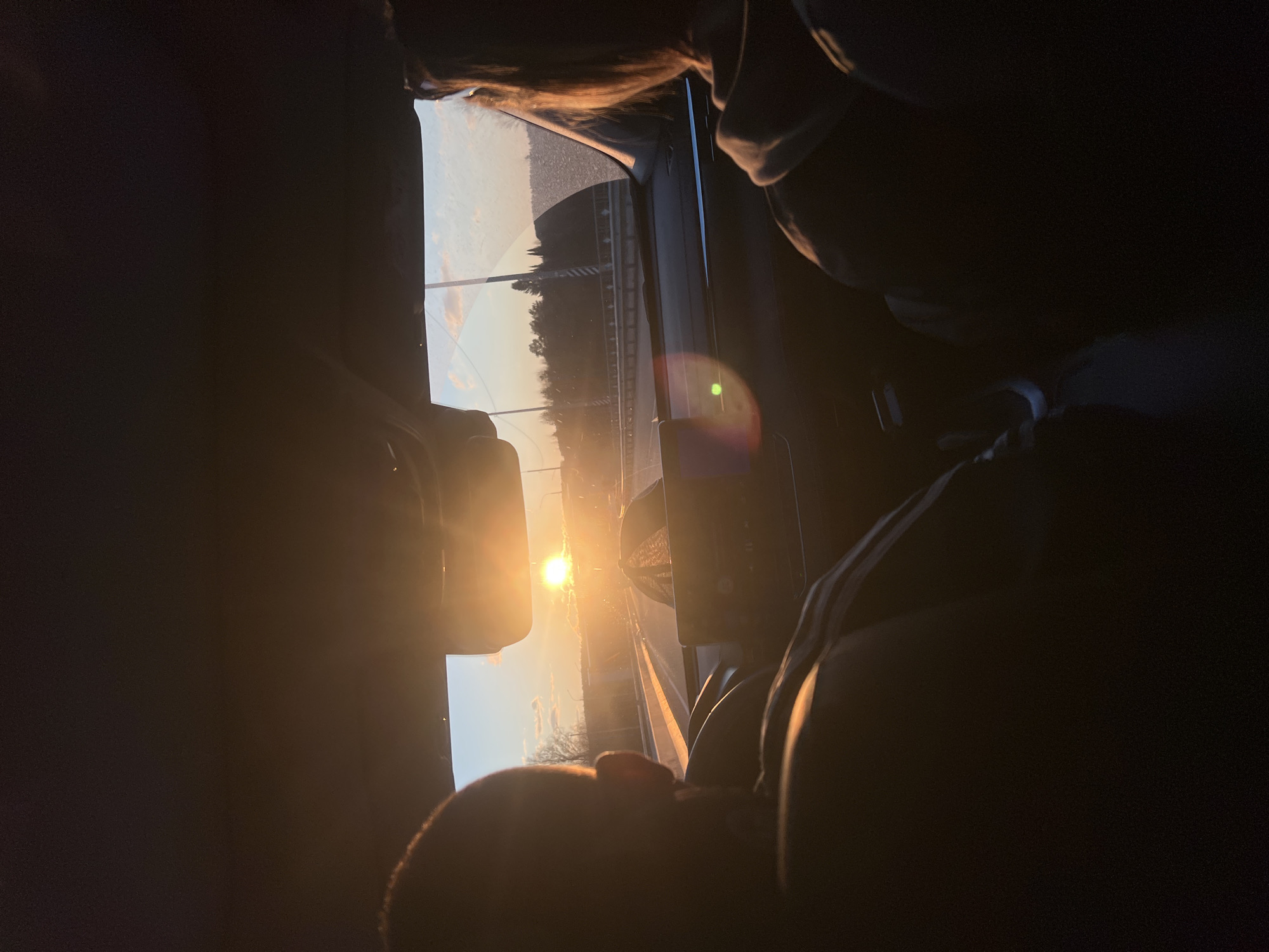 The image captures a dramatic sunset or sunrise viewed from inside a vehicle, likely a car or bus, in Tiush, Russia. The sun, a brilliant golden orb, is low on the horizon, intensely backlighting the scene and creating strong silhouettes within the vehicle.
In the foreground, parts of the vehicle's dark interior are visible, including what appears to be a dashboard or door frame on the left, and seats or passenger areas in the upper and right portions of the frame. Silhouetted against the bright light, a person's head is visible in the lower left, while another person's hair and upper body are seen towards the top, illuminated by the sun's glow.
Through the window, a road or highway can be discerned, bordered by what appears to be guardrails or barriers. Beyond the road, a line of distant trees is silhouetted against the bright sky. The sky itself shows warm hues typical of dawn or dusk, transitioning from bright gold near the sun to softer, lighter tones higher up, with a few faint clouds. A prominent green-blue lens flare is visible near the center-right of the image, a common effect when shooting directly into bright light.
The scene conveys the experience of traveling during the golden hour, with the sun dominating the outdoor view and casting deep shadows inside the moving vehicle. No visible text is present in the image.