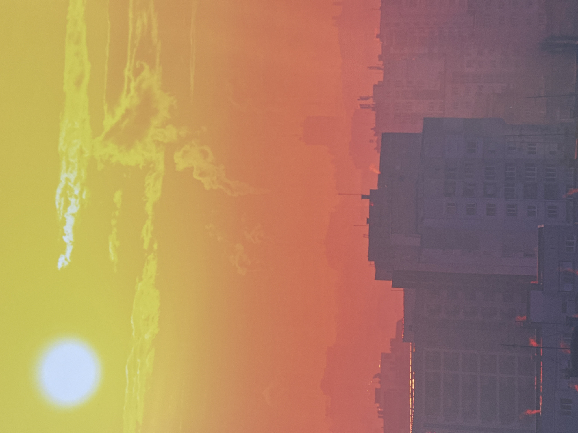 The image depicts a sunset over a city skyline, likely Sofia, Bulgaria. The left side of the image shows a bright sun, with streaks of clouds and light. The right side depicts dense buildings, appearing in a hazy purple hue. The sky transitions from bright yellow to orange to red, suggesting a sunset atmosphere. The buildings appear rectangular with multiple windows. No people are visible.