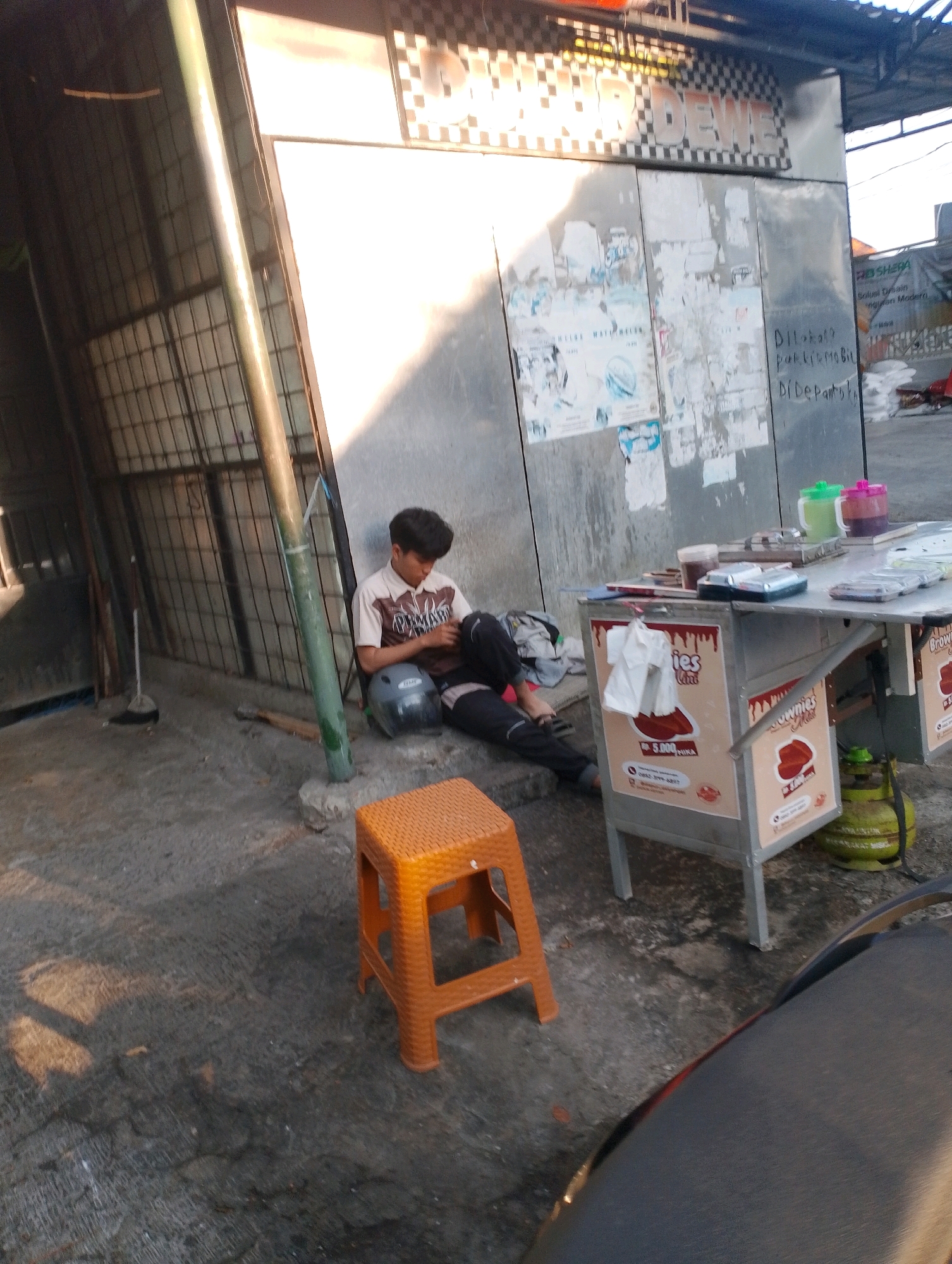 A young man is sitting on the ground, leaning against a grey metal structure, in an outdoor urban setting in Probolinggo, Indonesia. He is wearing a white and brown collared shirt and dark pants, and has a helmet on his lap. He appears to be interacting with a smartphone in his hands. In front of him is an orange plastic stool. To his right, a small food cart is set up, displaying advertisements for "Brownies". On the cart are several items, including colorful plastic cups, a container of food, and a silver electronic device. Behind the cart is a green gas cylinder. The ground is paved with concrete, and sunlight casts shadows on it. The metal structure behind the man has several posters or flyers attached to it. The overall scene suggests a street-side vendor area, possibly during a slow period.