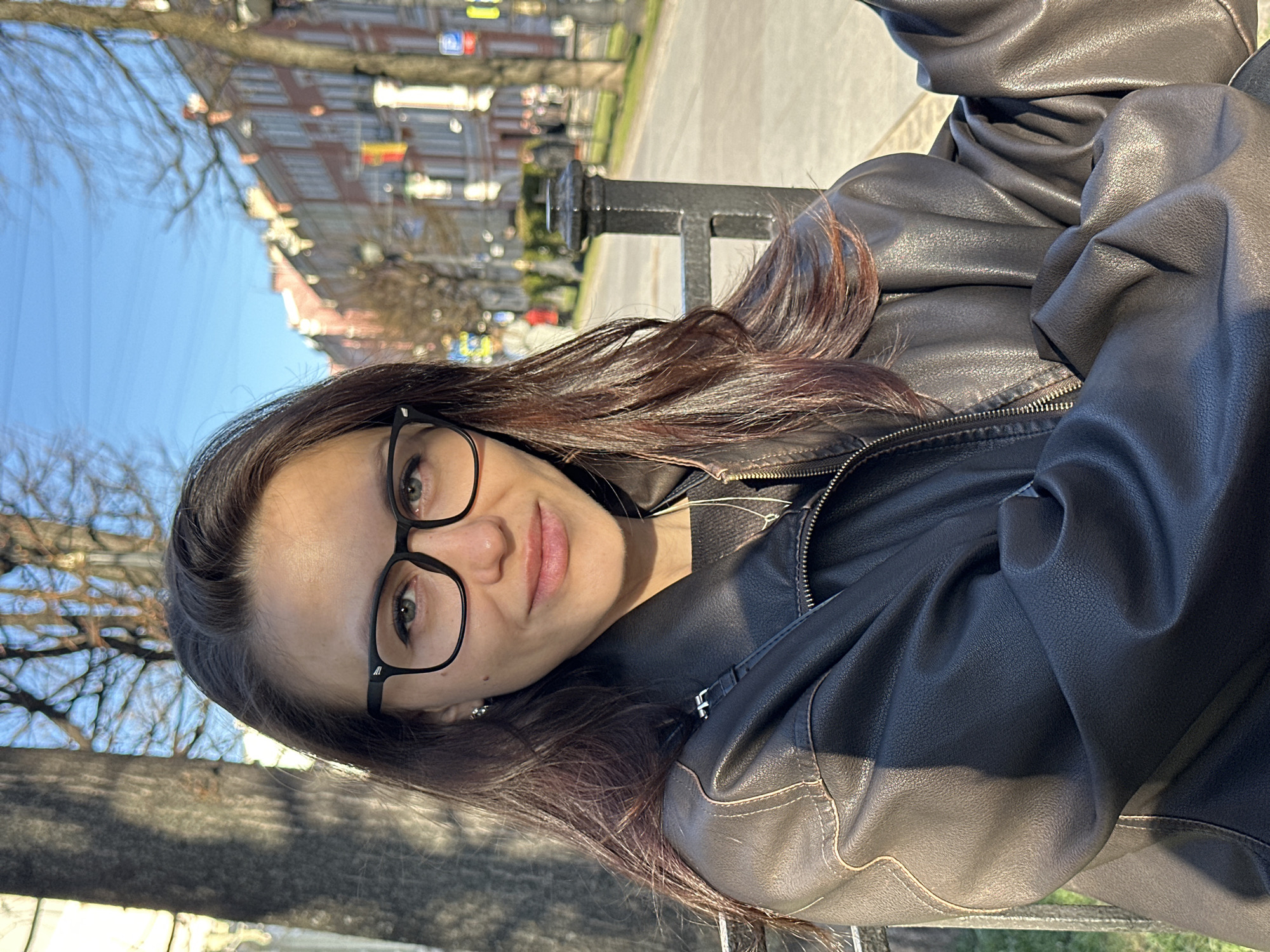 A woman with brown hair and glasses is posing for a photo in Saint Petersburg, Russia. She is wearing a black leather jacket and is sitting on a bench in a park. In the background, there are buildings and trees. The sky is blue and there is a streetlight on the corner.