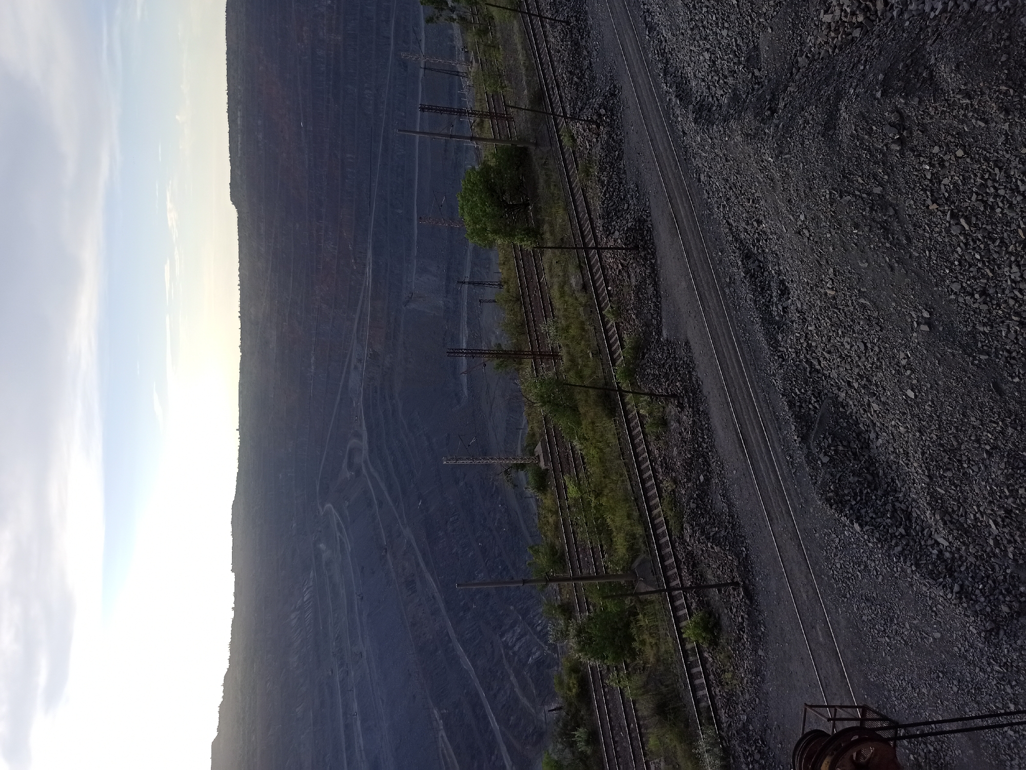 This media file depicts an aerial view of a large-scale mining operation, likely an open-pit mine. The scene is dominated by the vast, terraced slopes of the quarry, carved deep into the earth. The dark, layered rock formations suggest a significant excavation.
In the foreground, multiple sets of railroad tracks run parallel to the edge of the mine. These tracks are bordered by piles of dark, coarse aggregate, characteristic of mining waste or extracted material. Scattered vegetation, including trees and grass, grows along the edges of the tracks and in some areas within the mine itself, indicating a temperate climate and possibly a transition to a less active period of extraction.
Several utility poles with overhead wires are visible along the tracks, hinting at the operational infrastructure supporting the mine. At the very bottom of the frame, a piece of industrial equipment with visible gears and a rusty metal frame suggests ongoing or recent activity.
The sky, visible on the left side of the image, is bright with a mix of white and pale blue, indicating daylight, possibly early morning or late afternoon given the soft illumination. There is no visible text in the image. The overall impression is one of a massive industrial undertaking, characterized by scale and raw materials.