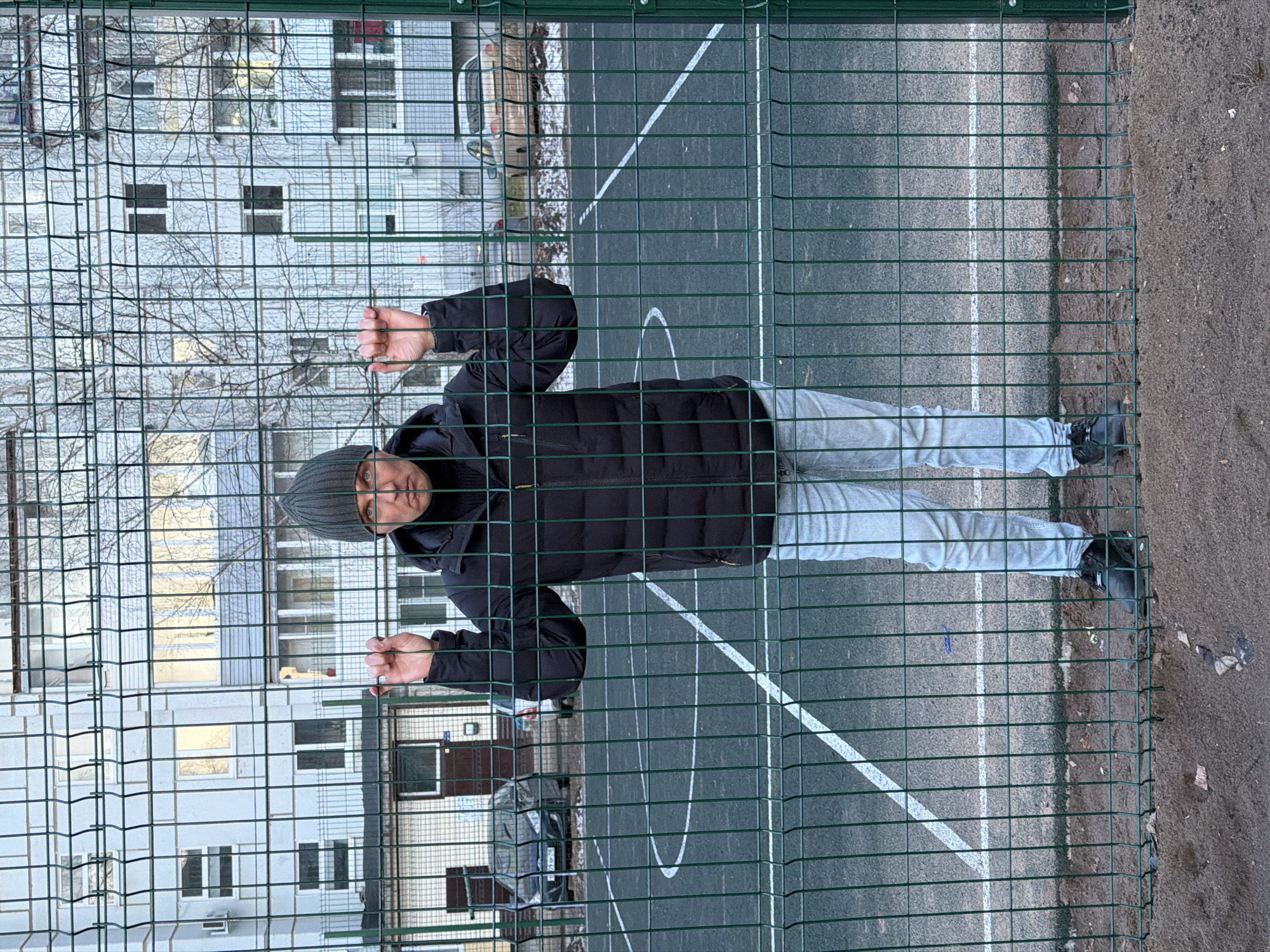 The image captures a male individual standing behind a green wire mesh fence on an outdoor sports court in Chaikovsky, Russia. The person is dressed in a dark puffer jacket, light grey pants, dark shoes, and a grey knitted beanie. He is seen gripping the fence with both hands, looking directly at the camera with a neutral expression.
The green fence, characterized by its rectangular grid pattern, dominates the foreground. Behind the fence lies a sports court with a dark asphalt or tarmac surface, marked with white lines, including a partial arc and straight boundary lines, indicative of a basketball or multi-purpose court. To the right of the court, a section of bare earth or sandy ground is visible.
In the background, beyond the court, stands a multi-story residential building, likely an apartment complex, featuring numerous windows. Bare trees, devoid of leaves, are positioned in front of the building, suggesting the season is late autumn, winter, or early spring. A dark vehicle is partially visible, parked near the building. The scene is illuminated by overcast daylight, consistent with the cool-weather attire and barren trees. No discernible text is clearly visible on any objects within the frame.