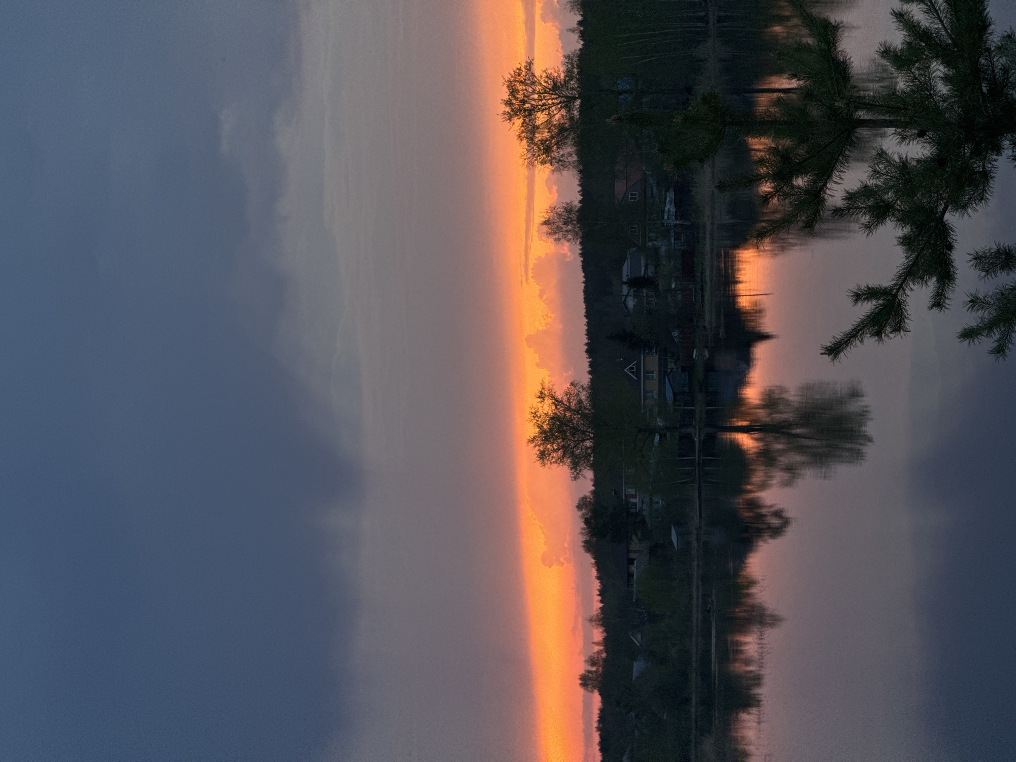The image depicts a tranquil scene at dusk in Bol'shoye Kozino, Russia. A body of water reflects the vibrant colors of the setting sun, creating a mirrored landscape. The horizon is ablaze with orange and pink hues, transitioning into a dark, cloudy sky. A line of trees and small houses sits along the water's edge, their silhouettes reflected in the calm surface. A pine tree branch extends into the frame, partially obscuring the view on the right. The overall atmosphere is peaceful and serene.