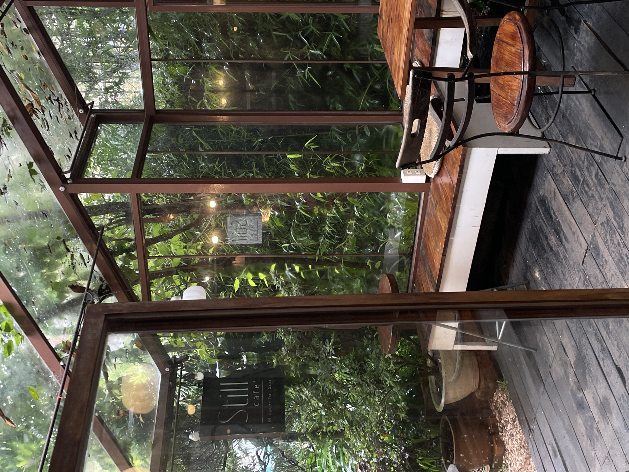 The image shows the interior of the "Still Cafe'" in Lâm Viên, Đà Lạt, Vietnam. The cafe appears to be enclosed in a glass structure, revealing dense greenery outside. Inside, there is wooden furniture, including a table and chairs, arranged on a wooden deck. The atmosphere seems tranquil. The sign for "Still cafe'" is visible through the glass, along with the address "59 Nguyen Trai, DaLat".