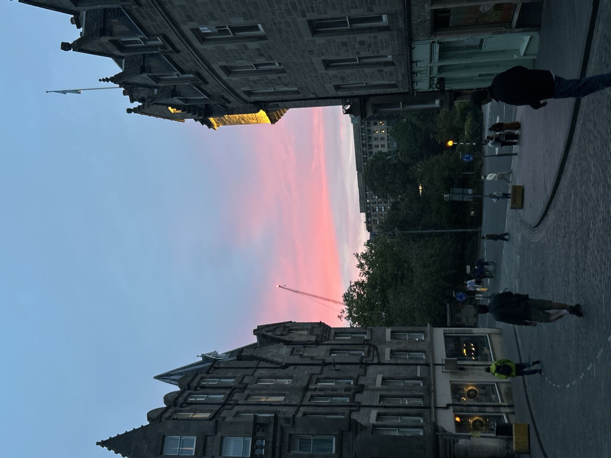 The image depicts a street scene in Edinburgh, Scotland, United Kingdom, during twilight. Two ornate stone buildings, characteristic of Edinburgh's architecture, flank a cobblestone street. The sky exhibits a gradient of blues and pinks, indicating the setting sun. A crane with red lights is visible in the distance, rising above trees. Several individuals are walking on the street, some appearing to be tourists. Shops are visible at the base of the buildings. The overall atmosphere is serene and captures the essence of an old European city at dusk.