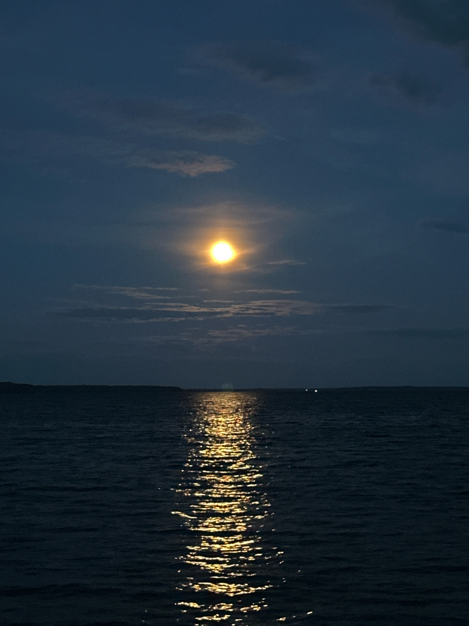 The image depicts a tranquil nighttime scene over a body of water, likely a lake or a wide river, near Нарацкі сельскі Савет, Belarus. The sky is a deep, dark blue, with scattered clouds that are illuminated by a bright, full moon. The moon, positioned slightly off-center in the upper portion of the frame, casts a strong, golden reflection onto the rippling surface of the water. The reflection creates a shimmering pathway of light leading towards the viewer. In the distance, along the horizon, a faint, dark line of land is visible, with a few distant lights suggesting the presence of human settlement or activity. The water itself is dark and appears somewhat choppy, with the moonlight creating highlights on the crests of the waves. The overall atmosphere is serene and peaceful, characteristic of a moonlit night in a rural or natural setting. There are no people or discernible animals visible in the image. The environment is dominated by the vast expanse of water and the illuminated night sky.