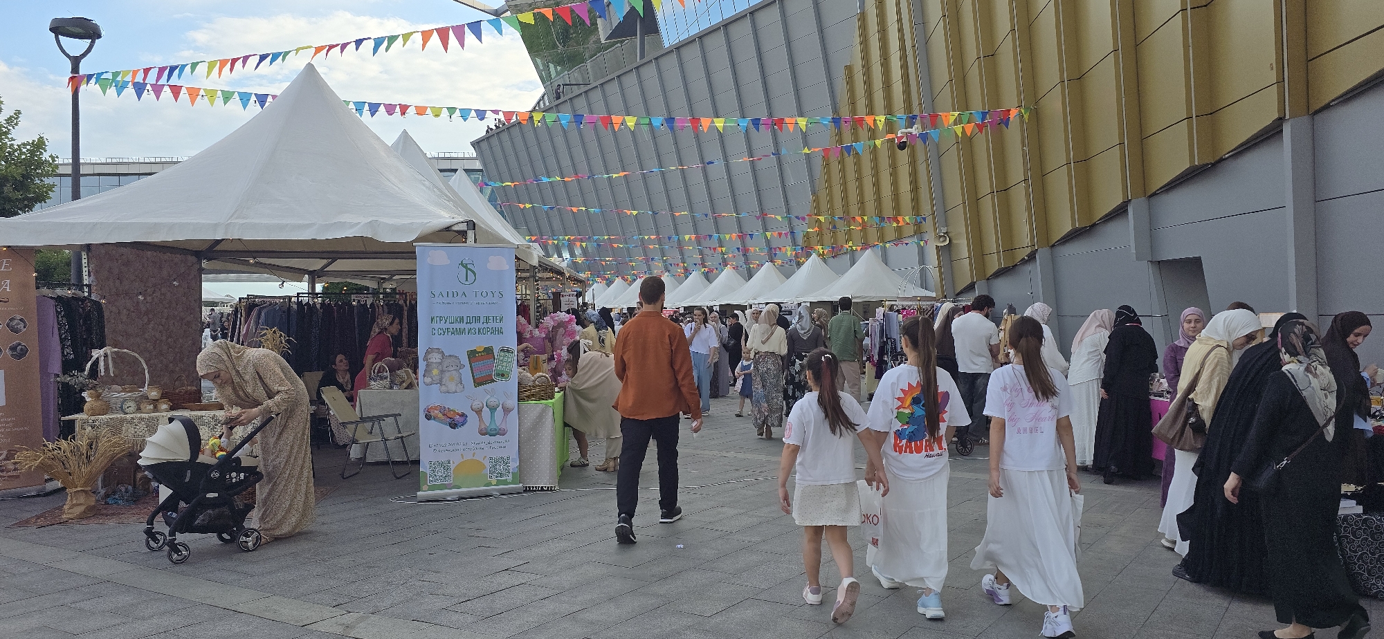 The image depicts an outdoor market scene in the city of Grozny, Russia, likely near the Grozny City shopping and entertainment center, given the distinctive architecture of the building in the background. The market features numerous white tents, suggesting a temporary setup for vendors. Colorful triangular flags are strung across the scene, adding a festive atmosphere. People of various ages and genders are present, many of whom appear to be of Muslim faith, with women wearing hijabs and abayas.
In the foreground, a woman in a beige, patterned hijab and dress is attending to a baby in a black stroller. Next to her, a table displays woven baskets, decorative items, and fabric samples. A large banner for "SAIDA TOYS" is prominently displayed, featuring images of children's toys and text in Russian.
Further into the market, a man in an orange shirt and black pants walks away from the camera. A group of young girls in white dresses are walking together, holding hands. Other individuals and groups are browsing stalls and interacting with vendors. The ground is paved with gray stones. The overall impression is a bustling community event, possibly a fair or festival, set against a backdrop of modern urban architecture.