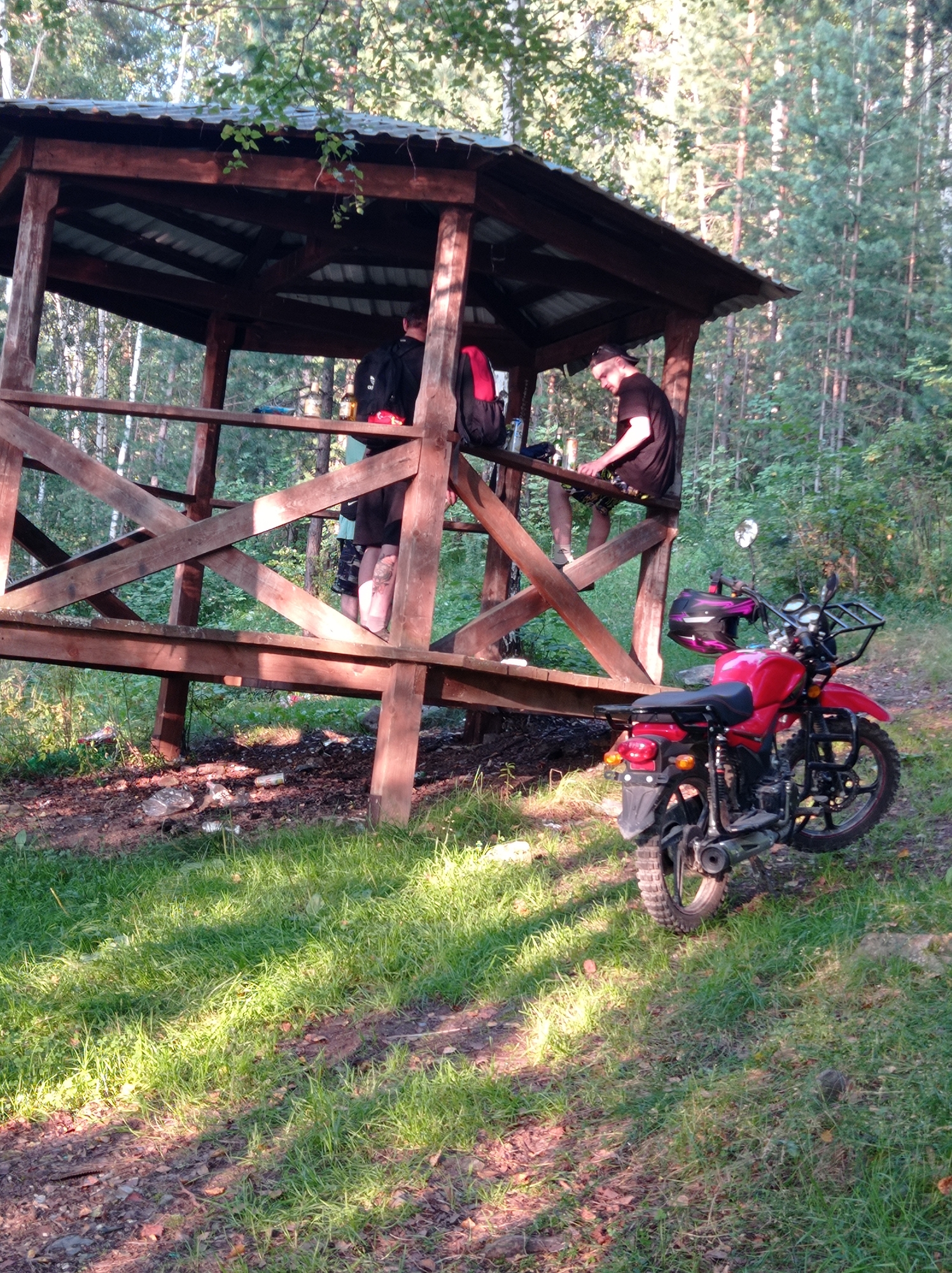 A red motorcycle is parked on a grassy area near a wooden gazebo in a forest setting. Two individuals are visible within the gazebo. One person, wearing a black t-shirt and a cap, is leaning on the railing and looking down at something on a table. Another person, wearing a black shirt and shorts, is standing with a backpack on their back. The ground around the gazebo is covered with grass and debris, including plastic bottles and wrappers. The sun is casting dappled shadows through the trees. The location is Degtyarsk, Russia.