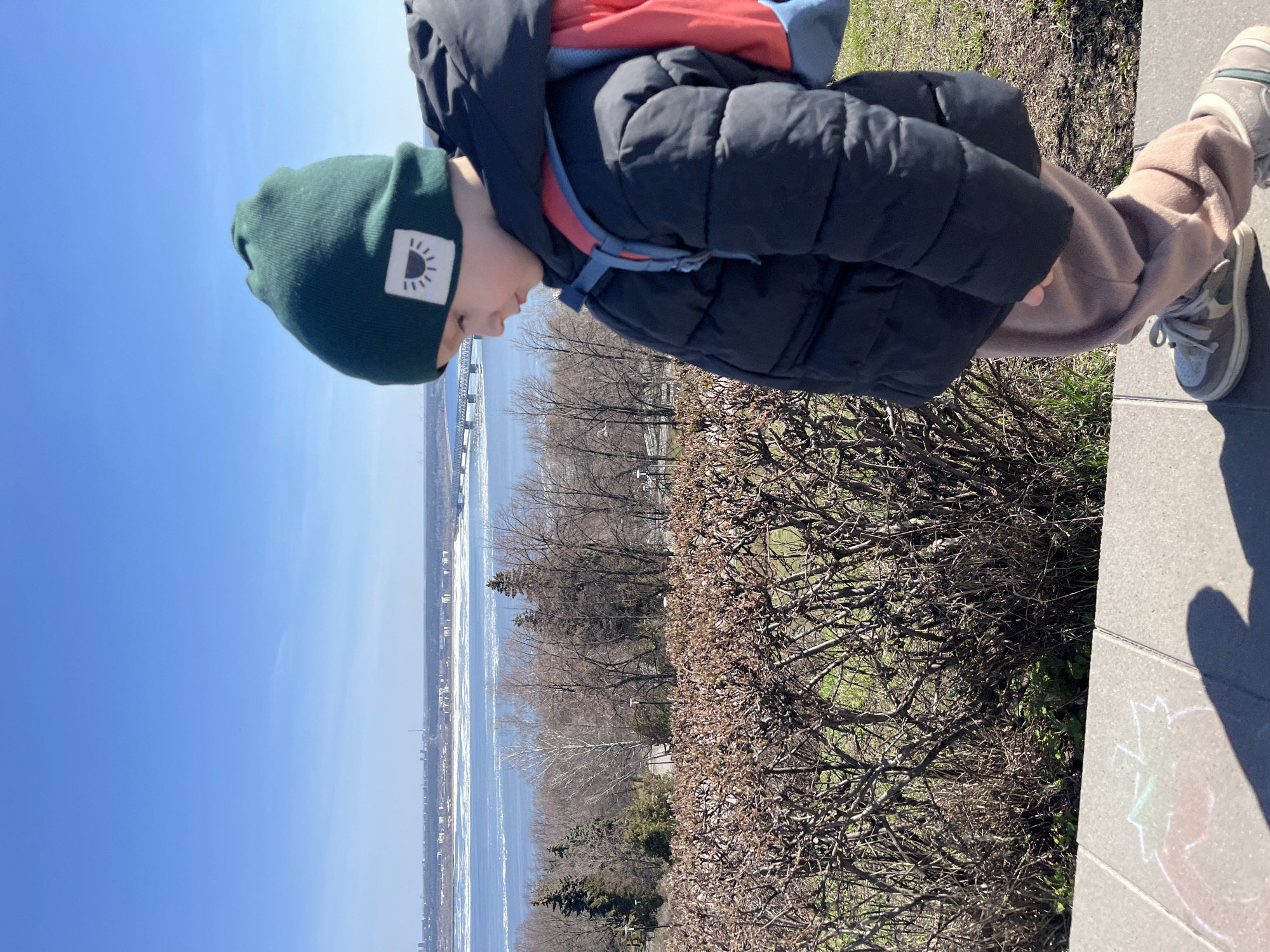 A young child wearing a green beanie, black puffer jacket, beige sweatpants, and grey shoes is standing on a concrete ledge in Ulyanovsk, Russia. The child is looking down towards a body of water, likely the Volga River, which is visible in the background. Beyond the water, a cityscape stretches along the horizon. In front of the child and the ledge are bushes and trees, some bare and some with sparse foliage. The sky is clear and blue. The image is shot from a low angle and is rotated counter-clockwise. Chalk markings are visible on the concrete ledge.