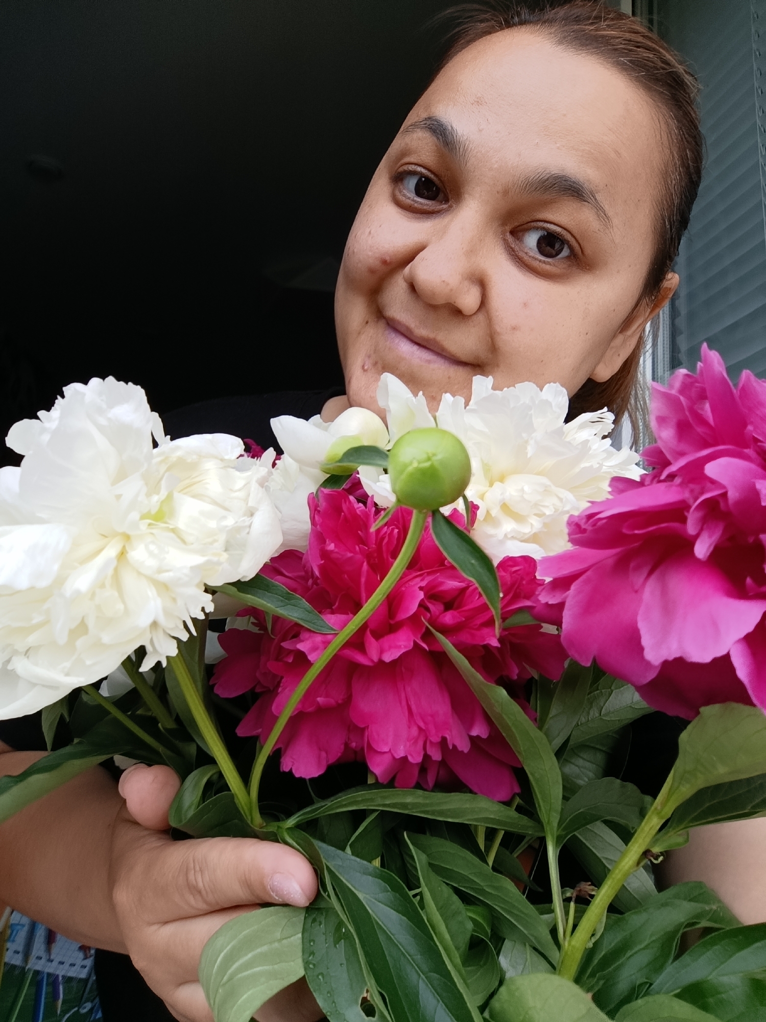 A medium shot captures a woman from the chest up, holding a bouquet of vibrant pink and white peonies. She is looking towards the camera with a soft smile and her head is tilted to the right. Her hair is brown and pulled back. The peonies are in full bloom and dominate the foreground, with a single green bud visible. The background is dark on the left and features a textured surface, possibly a window blind, on the right. The scene appears to be indoors. There is no indication of the city of Chaikovsky, Russia.