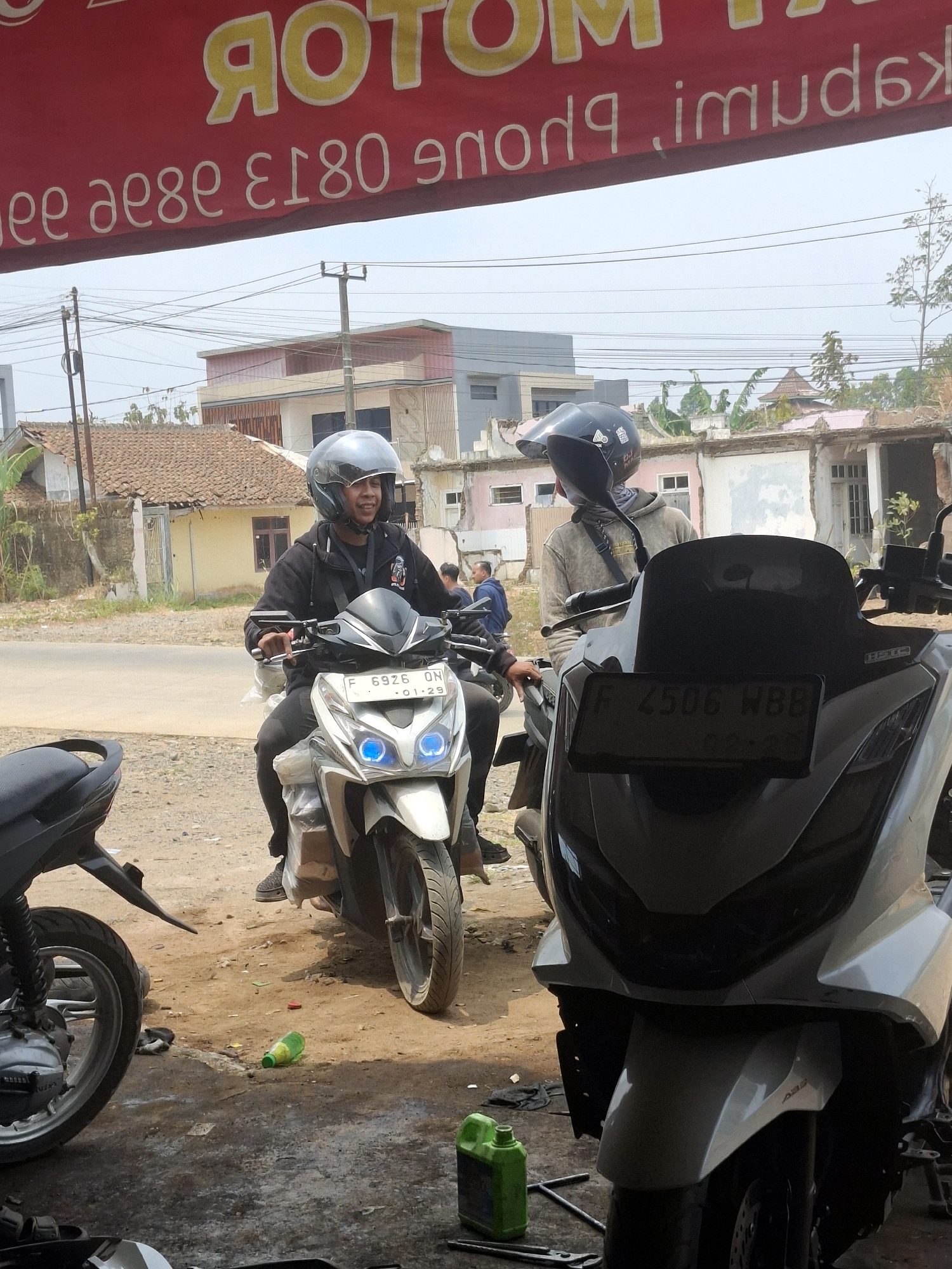 A man wearing a black hoodie and a silver helmet is riding a white motorcycle. He is looking to his right and smiling. Another man in a beige hoodie and a black helmet is standing next to him on another motorcycle. The motorcycles are parked on a dirt road in front of a building. There are other buildings and trees in the background. The scene appears to be a motorcycle repair shop. The license plate on the white motorcycle reads "F 6926 ON". The license plate on the other motorcycle reads "F 4506 WBB". The location is in Ciheulang Tonggoh, Indonesia.