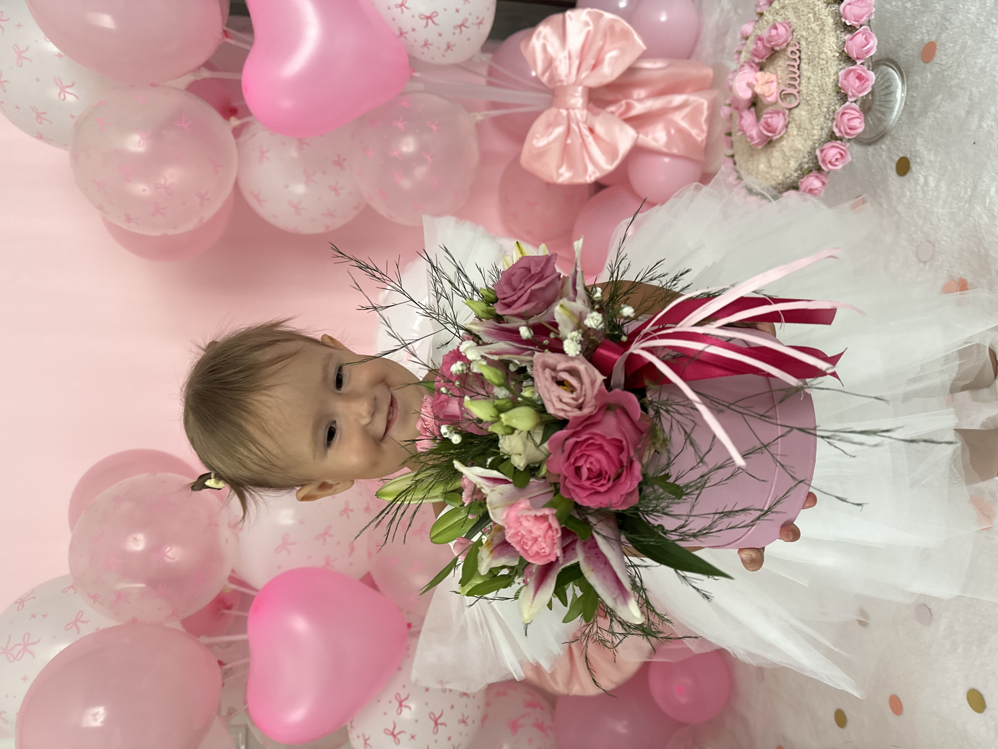 In a brightly lit indoor setting, a young girl with a ponytail and a delighted expression holds a pink flower box filled with a bouquet of pink and white flowers. She wears a white, frilly dress. The background is adorned with numerous pink and white balloons, some featuring bow patterns. To the right of the girl, a cake decorated with pink roses and the name "Olivia" is displayed on a glass stand. The scene suggests a birthday celebration. The image does not provide any information about the location in the city of Wisła, Poland.