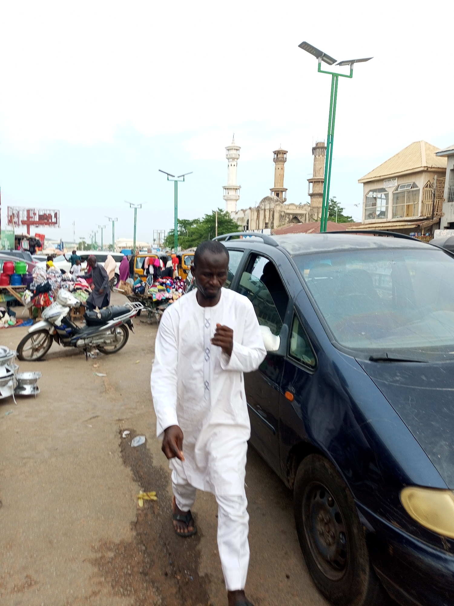 A man in a white tunic walks past a dark blue minivan in Arawa, Nigeria. He is walking from right to left, looking towards the viewer. The minivan is parked on the right side of the frame, with its hood and passenger-side door visible. In the background, a busy marketplace is set up with stalls selling various goods, including textiles and household items. Several motorcycles are parked in the market, and people are browsing the merchandise. In the distance, several minarets of a mosque can be seen, suggesting a religious and commercial hub within the city. The street appears to be paved with asphalt, and the sky is overcast, indicating a cloudy day.
