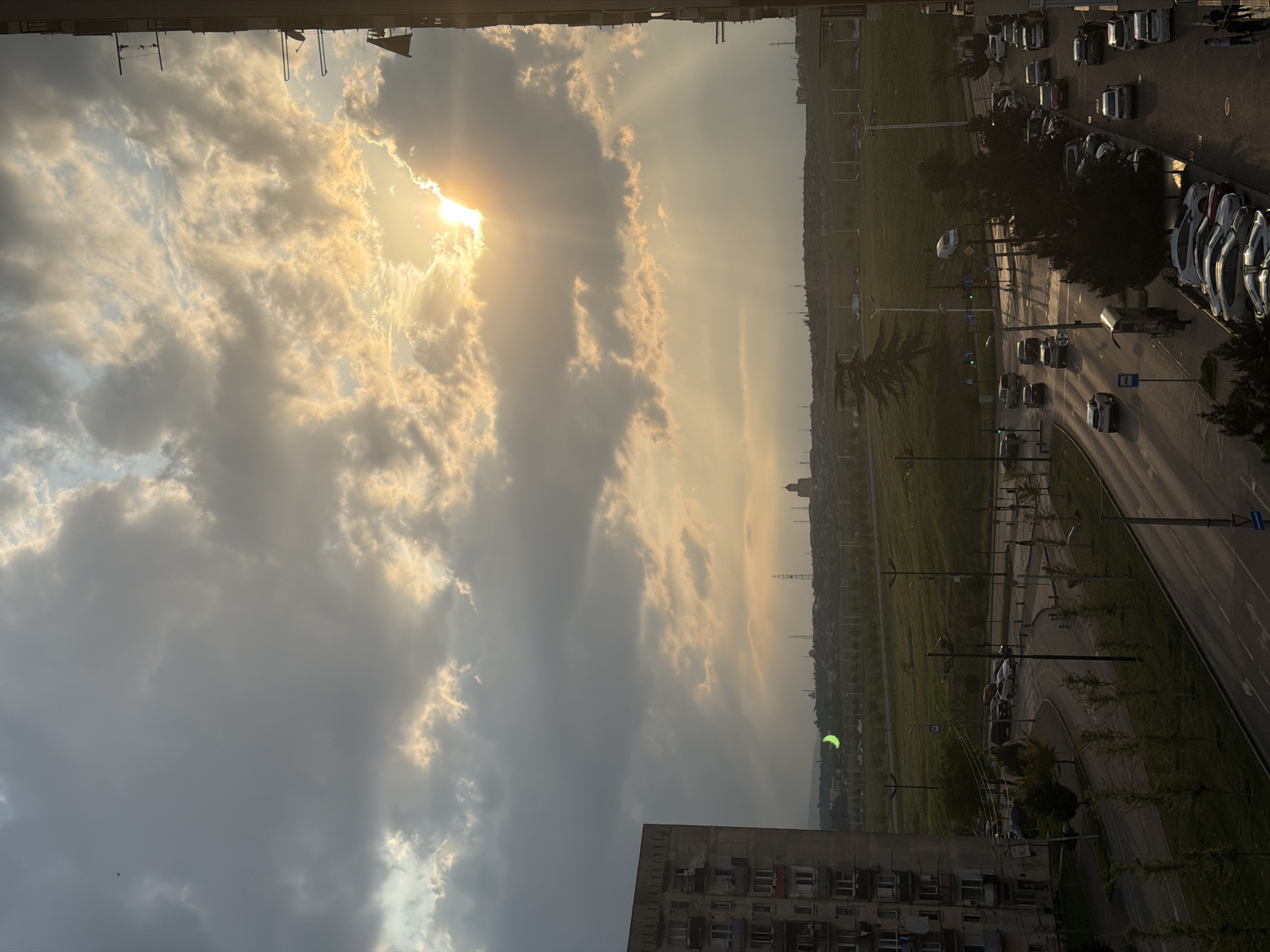 The image shows a view from a high vantage point, likely a building, overlooking a portion of Tbilisi, Georgia. The sky is dominated by clouds, with the sun visible through a break in the clouds, creating a bright, diffused light. Below the clouds, there is a cityscape consisting of residential buildings, a major roadway, green spaces, and distant urban structures. The roadway appears to have vehicular traffic, with cars visible. Streetlights line the road. The overall impression is that of a city scene under a dramatic sky with the sun positioned behind the clouds.
