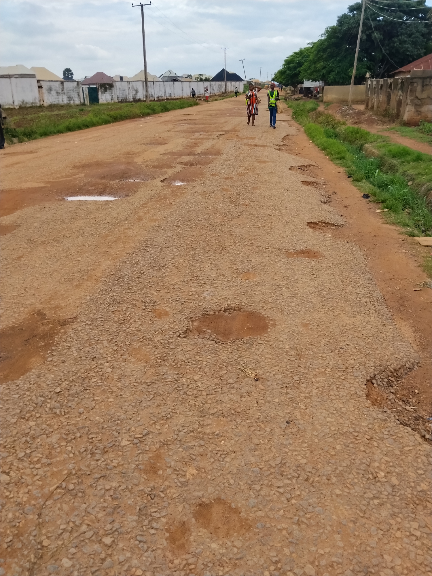 The image depicts a rough, unpaved road in Mangu Town, Nigeria. The road surface is uneven and filled with numerous potholes, some containing stagnant water. The surrounding environment includes grassy areas and a line of low buildings with white walls and various roofing materials. Two individuals are walking on the road; one, a woman wearing a striped dress, is pointing ahead, and the other, a person in a yellow safety vest and blue trousers, is walking beside her. In the background, utility poles and trees are visible. The sky is overcast.
